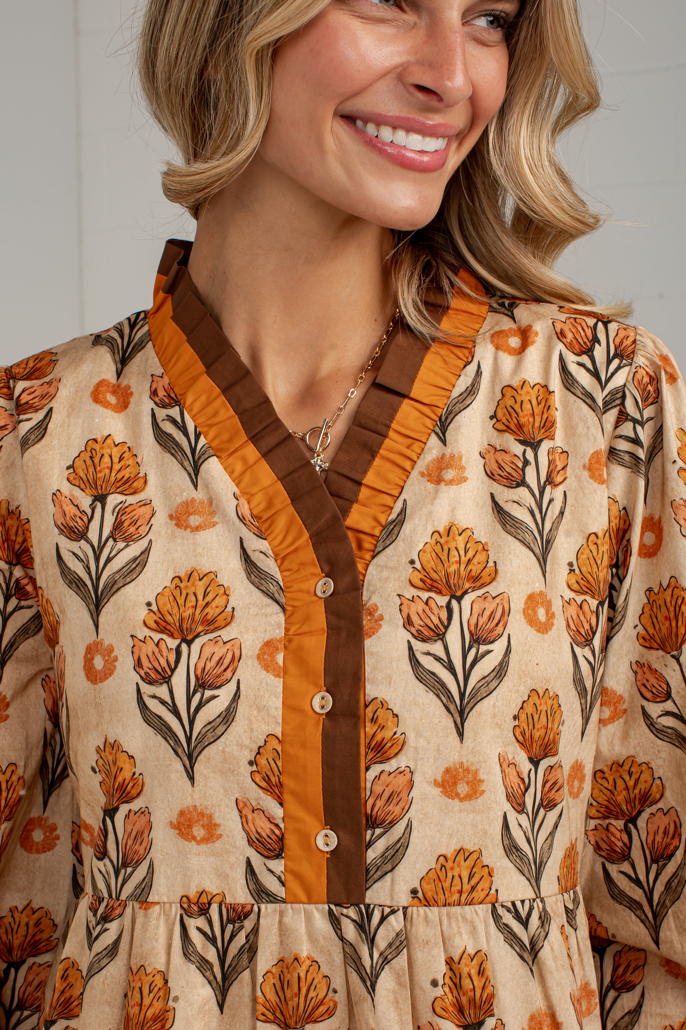 A close-up of a woman wearing a beige long-sleeve blouse with an orange and brown floral print, featuring a ruffled V-neckline and a brown button placket.
