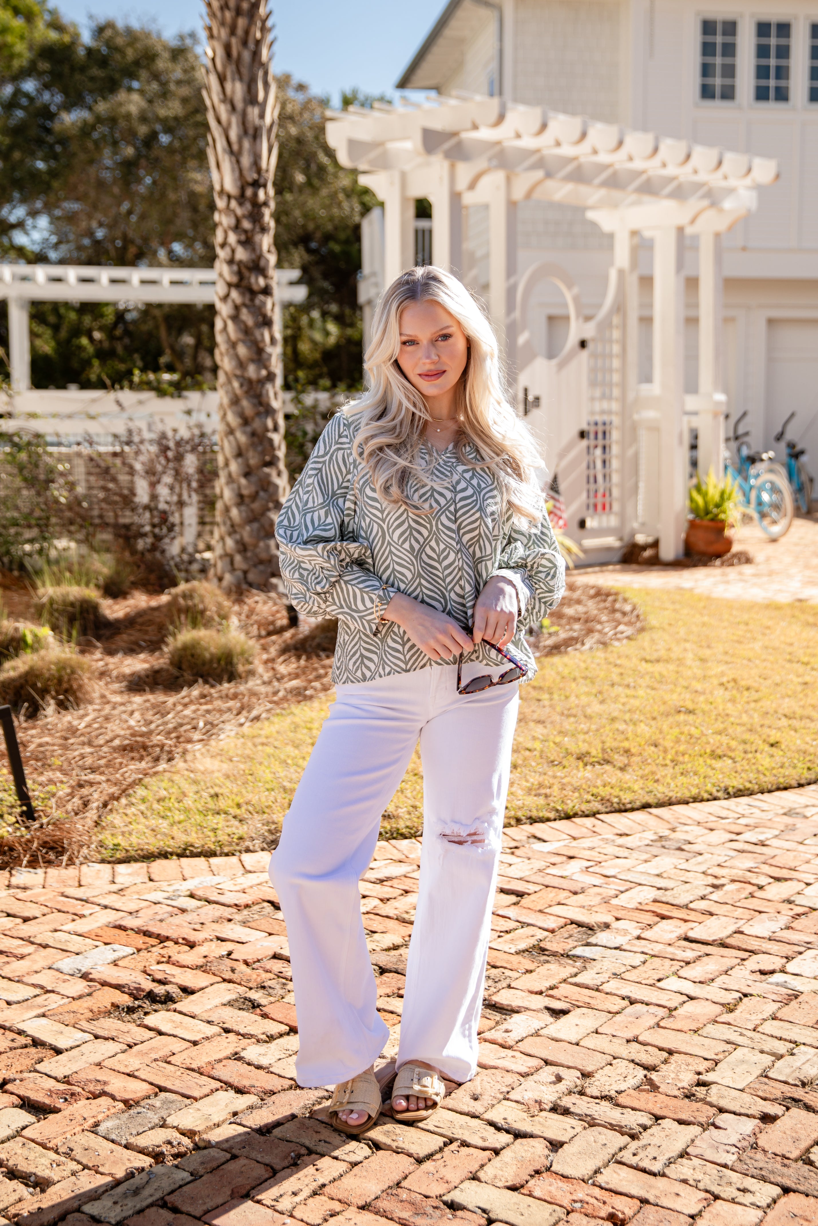 Woman standing on a brick patio in a garden setting with a house and pergola in the background.