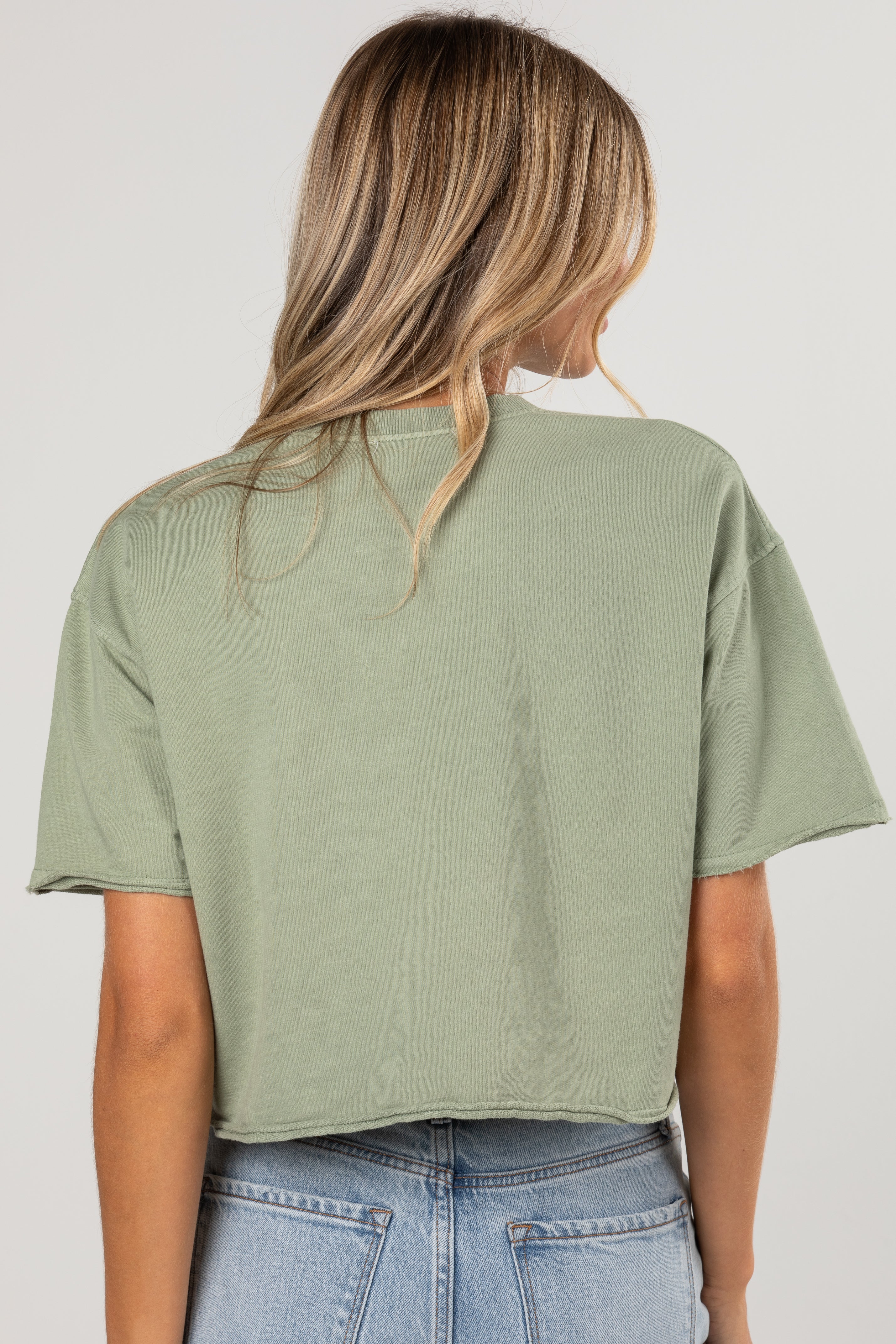 A woman viewed from the back wearing an oversized sage green short-sleeve crop top and light blue jeans against a plain light gray studio background.
