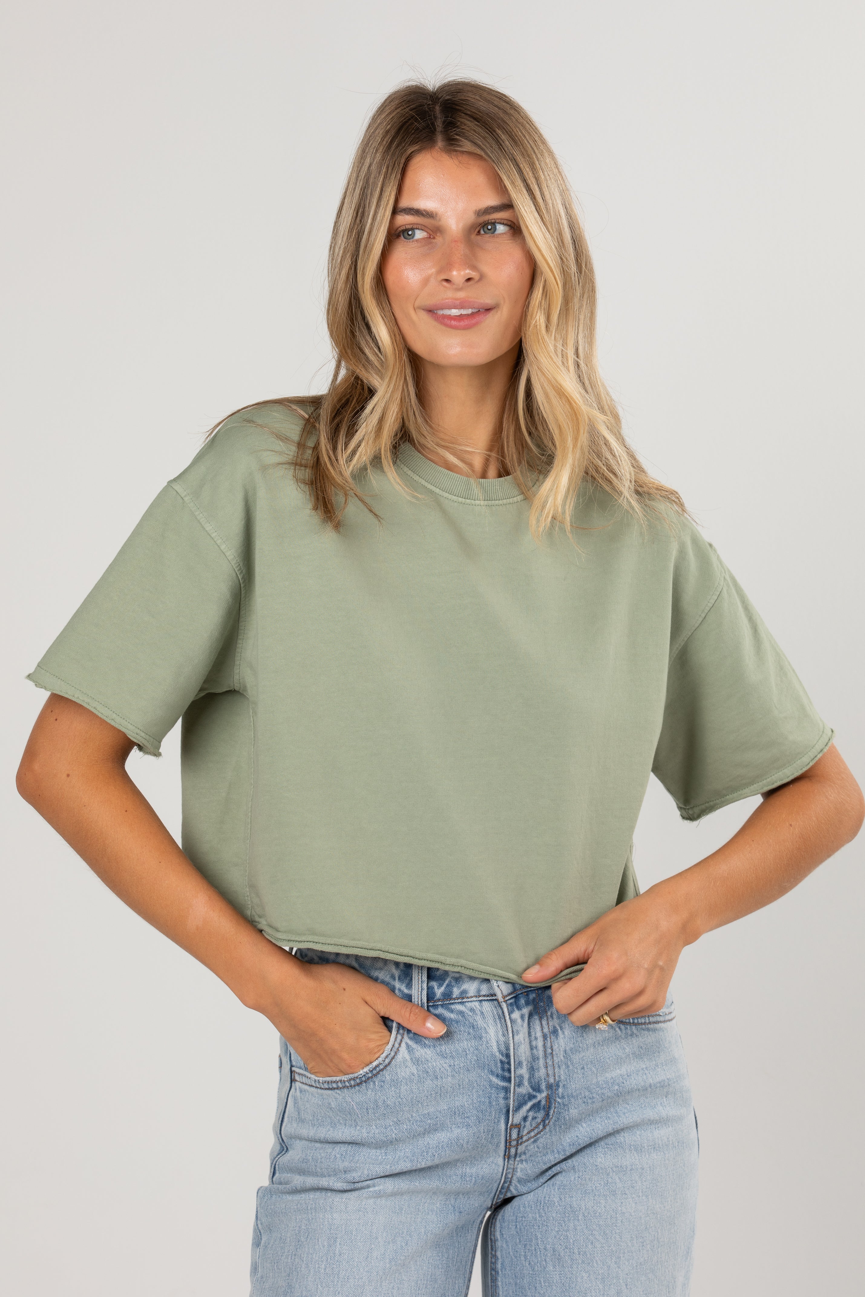 A woman wearing a light sage green short-sleeve crop top and light blue jeans, standing against a neutral light gray studio background.