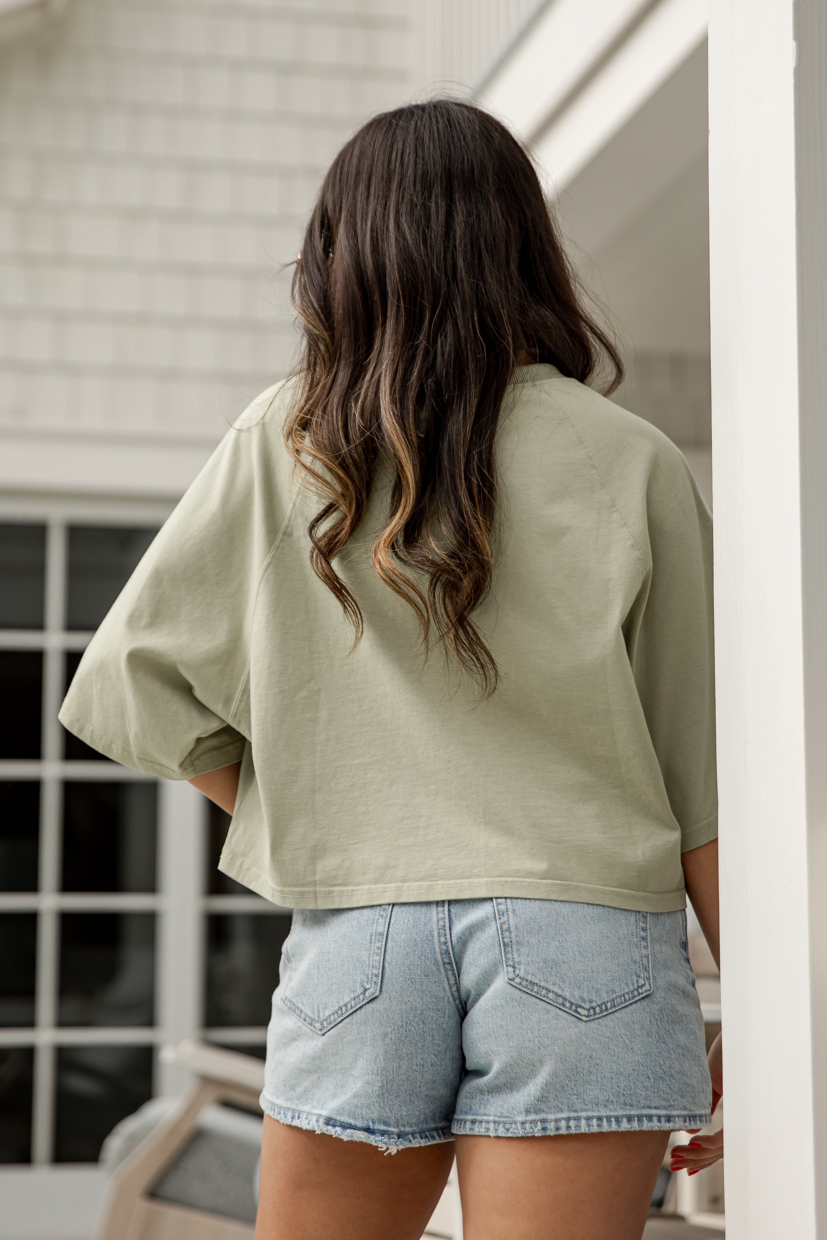 Person wearing a light green oversized shirt and denim shorts standing in a room with a window.