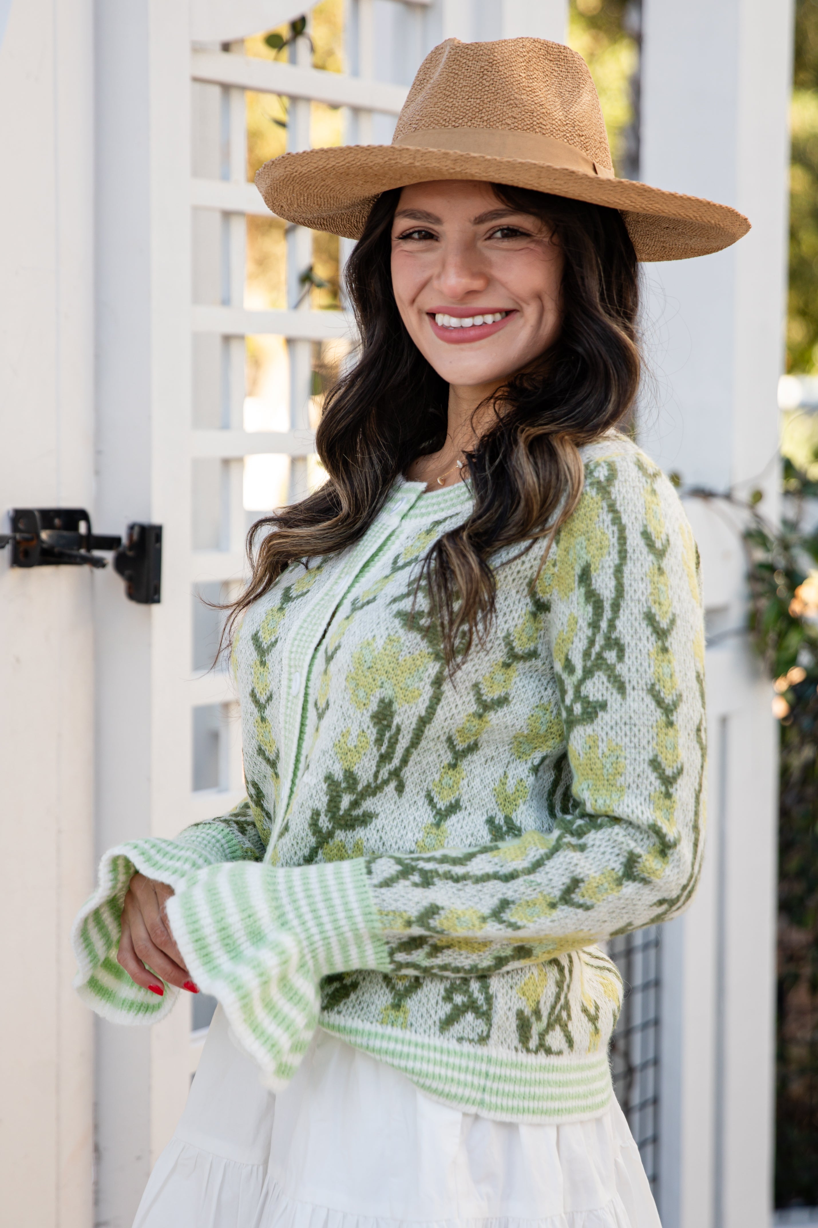 Woman wearing a green patterned sweater and brown hat standing in front of a white lattice.
