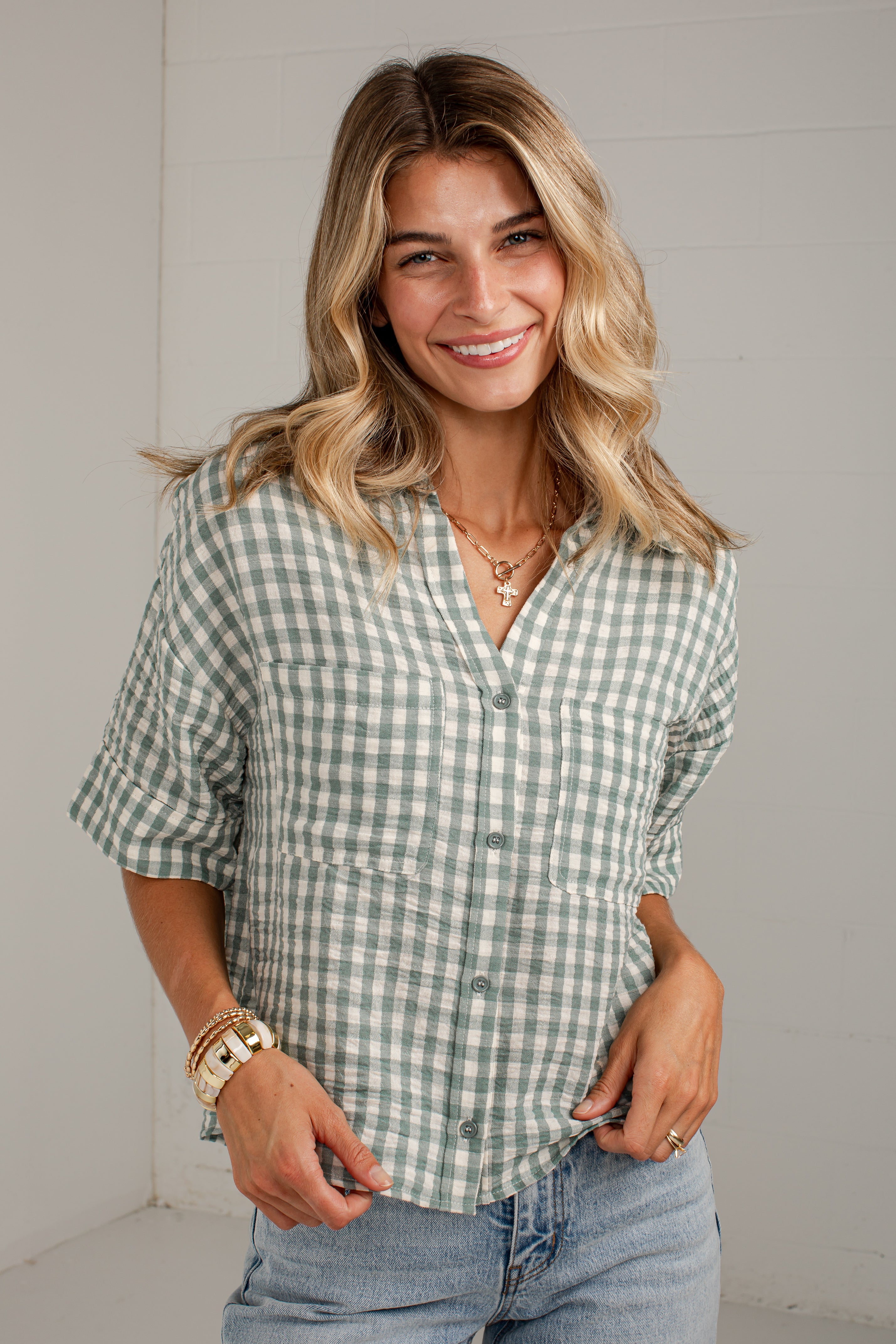 A smiling blonde woman wearing a sage green and white gingham short-sleeve button-up shirt with rolled cuffs, paired with light-wash blue jeans, standing against a white paneled wall.