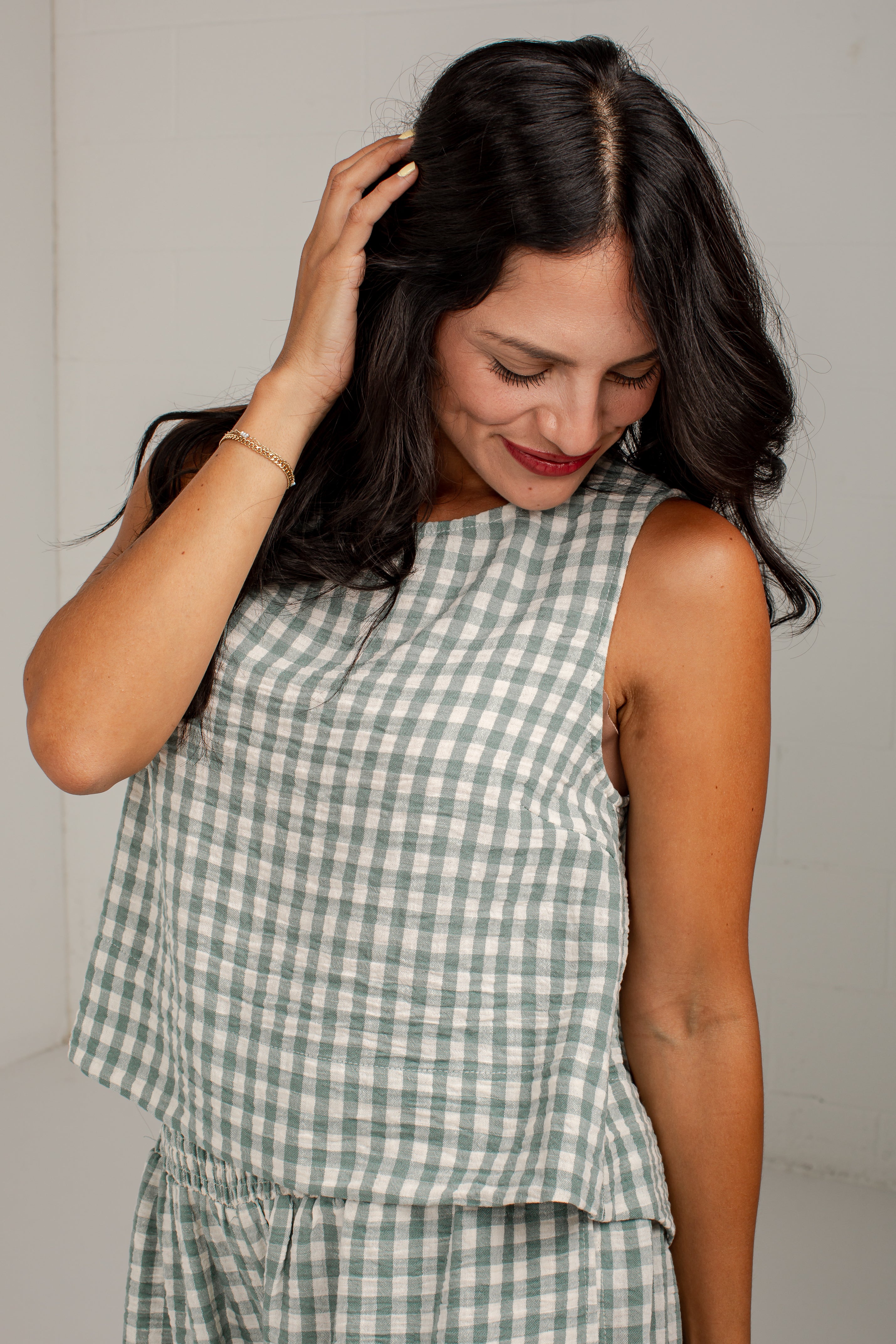 A woman wearing a sleeveless green and white gingham check top, shown from the chest down against a plain light gray wall.