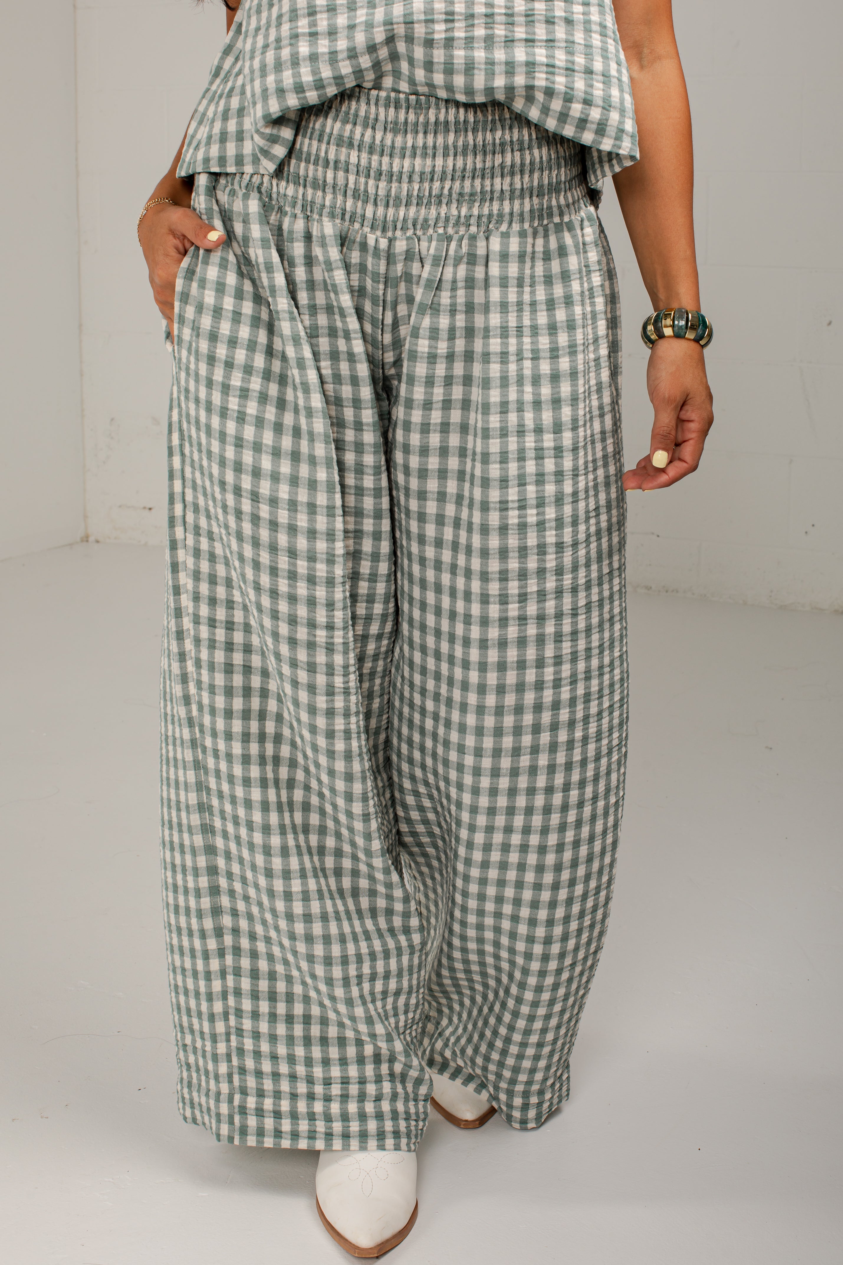 A person wearing wide-leg cropped pants in a green and white gingham check pattern, paired with a matching green and white gingham top and white pointed-toe heels, photographed against a plain light gray studio background.