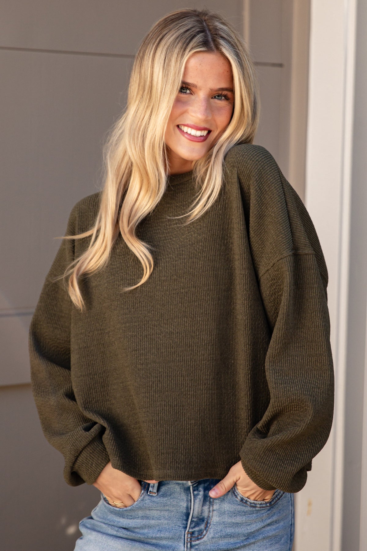 A smiling blonde woman wearing an oversized olive green knit sweater and light blue jeans, standing in front of a beige garage door and white brick wall.
