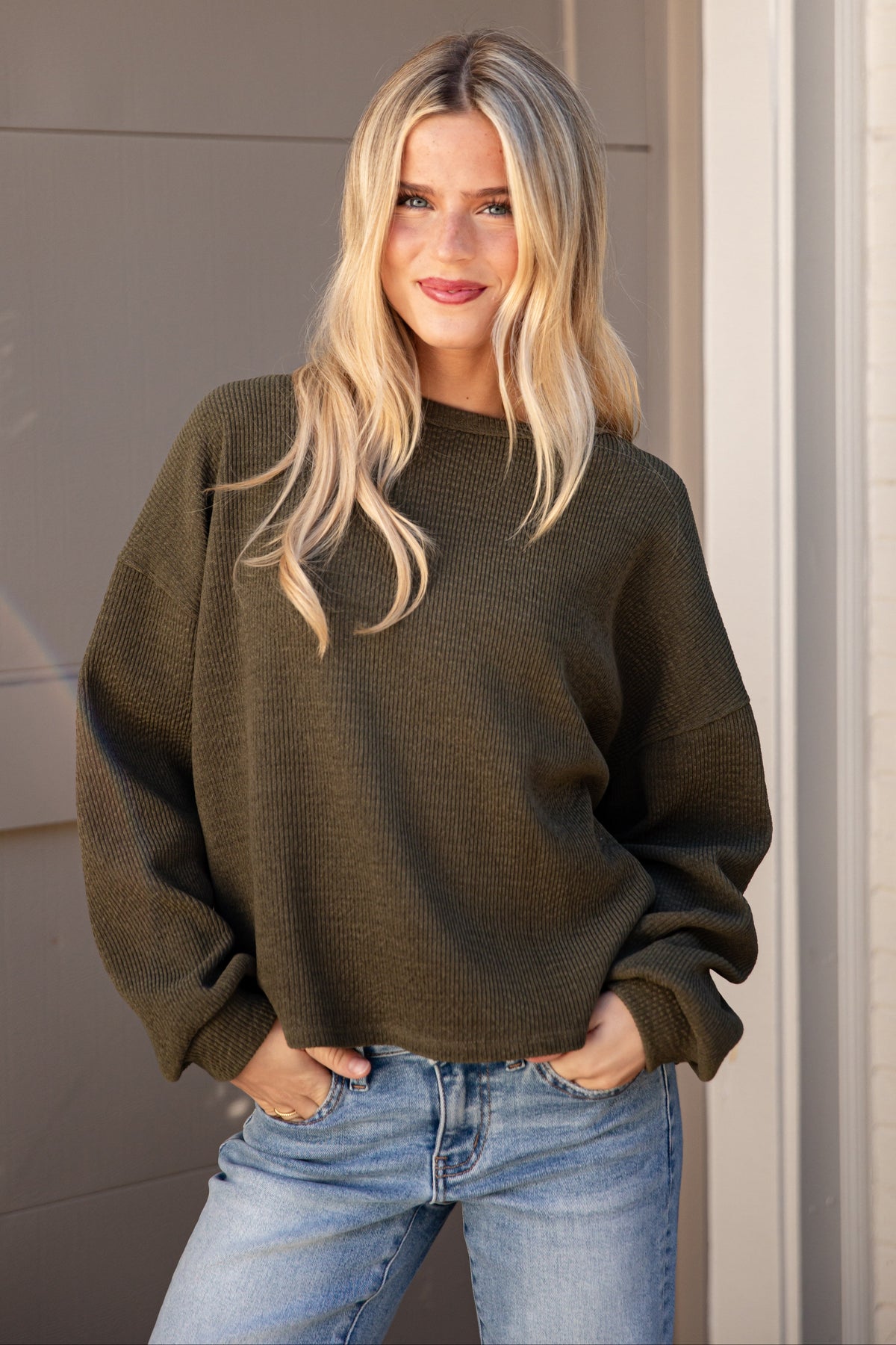 A woman with blonde hair wearing an olive green knit crewneck sweater and light blue jeans, standing in front of a beige garage door.