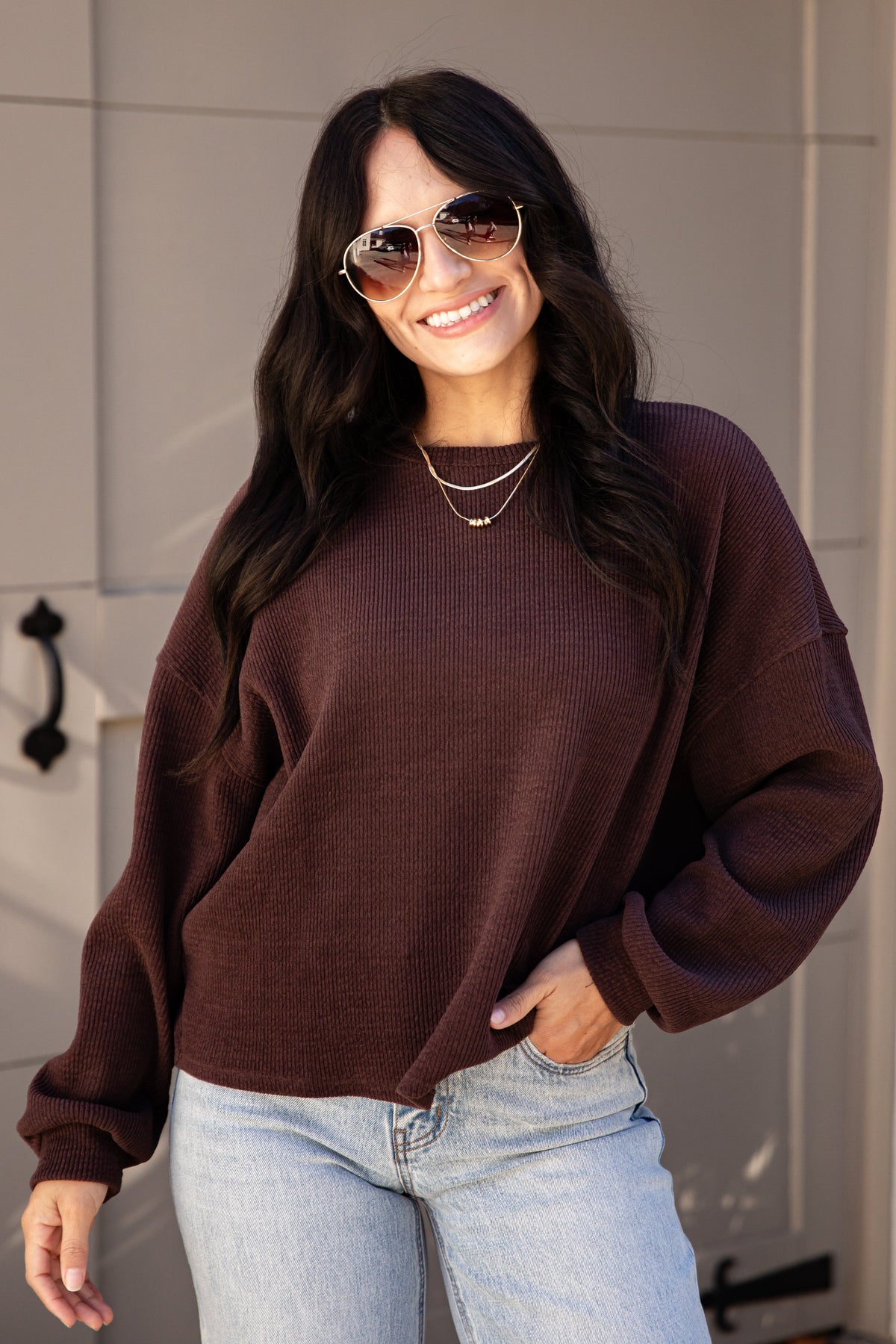 A woman wearing a dark brown waffle-knit long-sleeve sweater and light blue jeans, standing in front of a modern paneled wall.