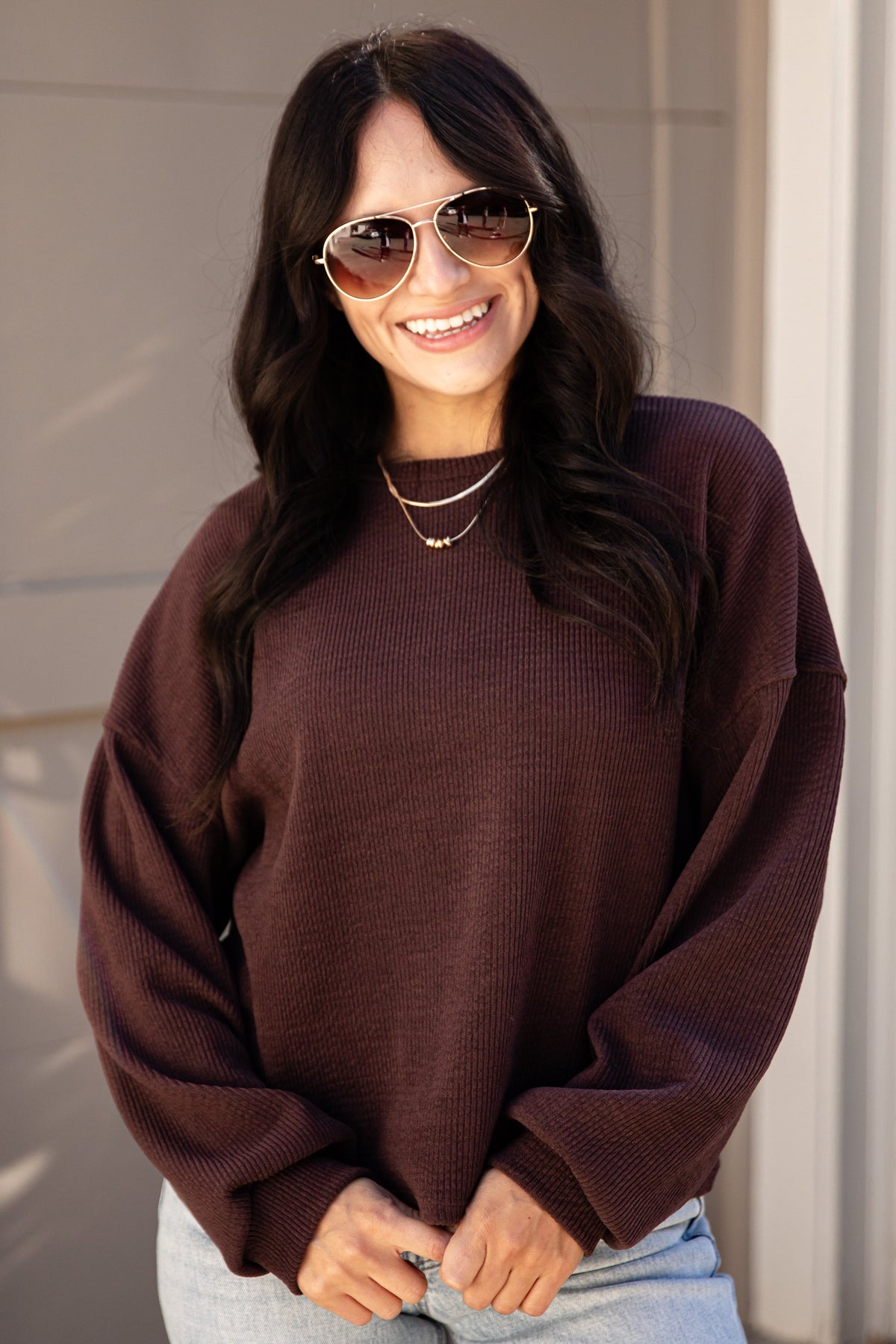 A woman wearing a dark brown waffle-knit crewneck sweater and light blue jeans, standing outdoors in front of a beige paneled door.