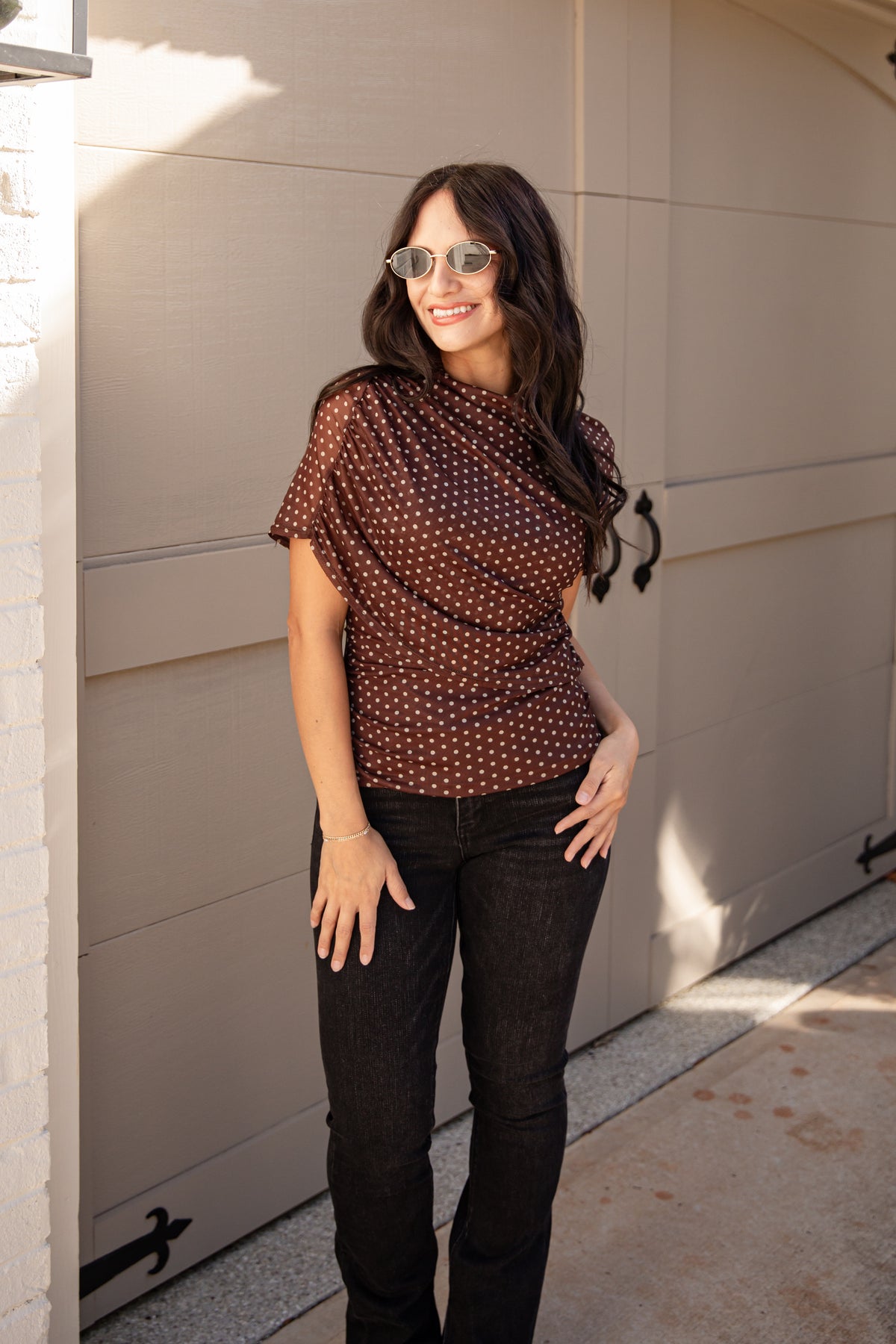 A woman wearing a brown short-sleeve top with a white polka-dot pattern and black jeans, standing in front of a beige garage door.