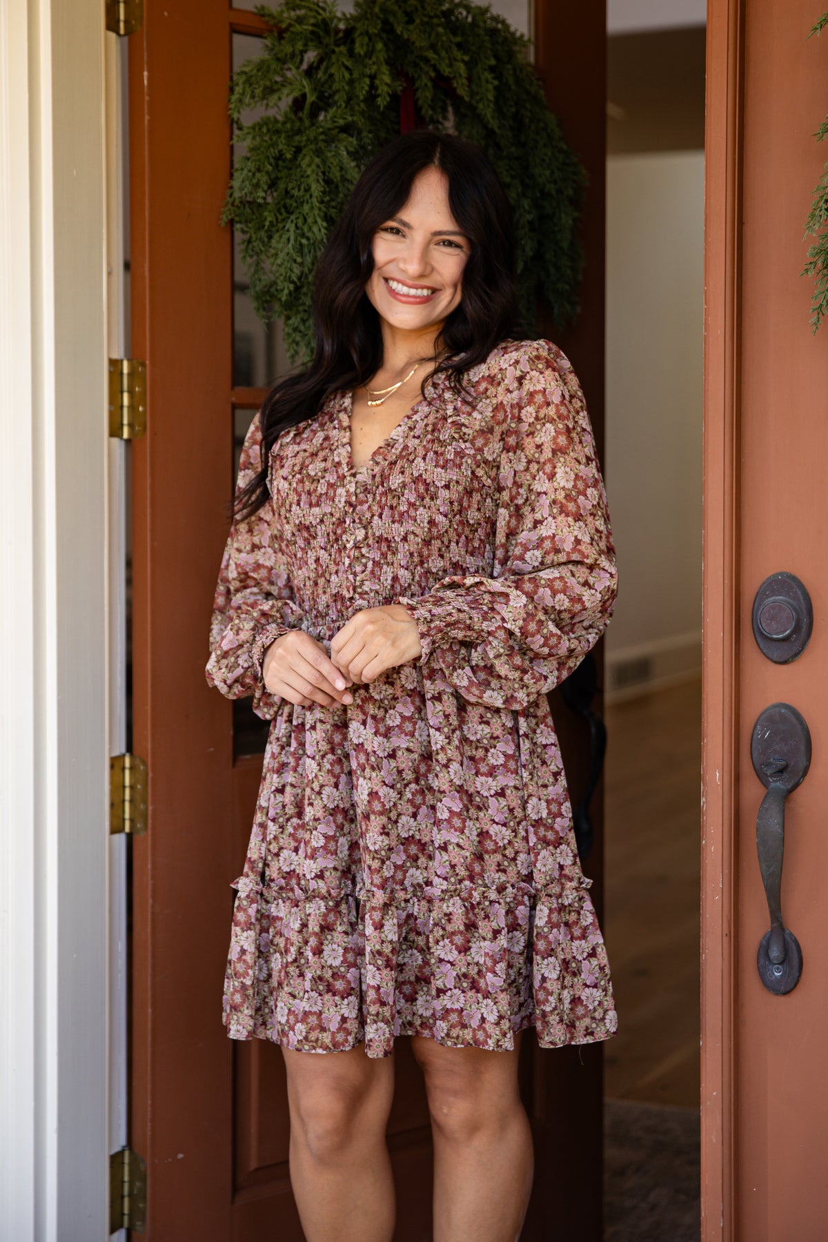 A woman stands smiling in a doorway wearing a long-sleeve floral mini dress with a wrap-style V-neck and tie at the waist. The dress has a muted pink, brown, and beige floral pattern.