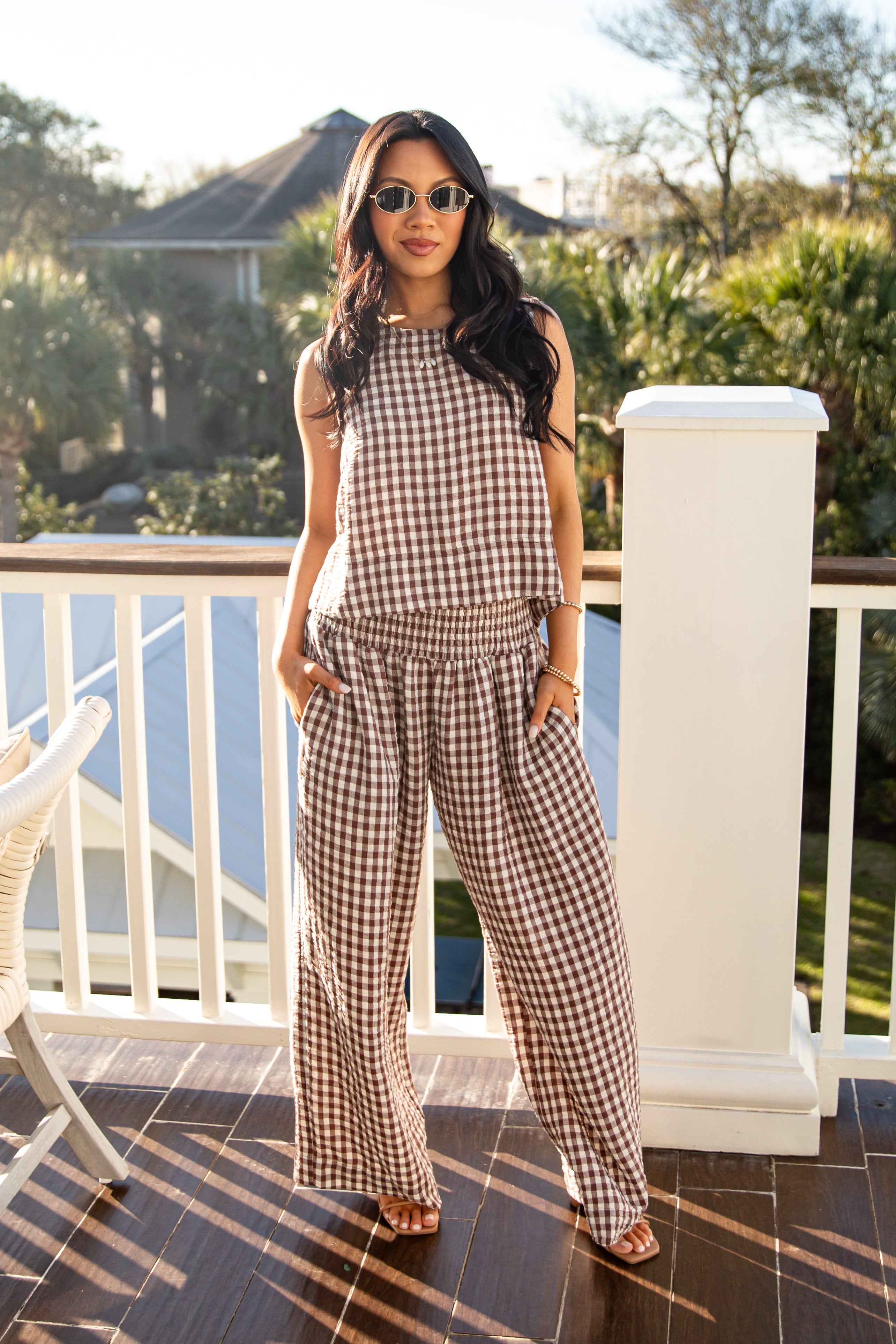 Woman in a checkered outfit standing on a balcony with trees and a house in the background.