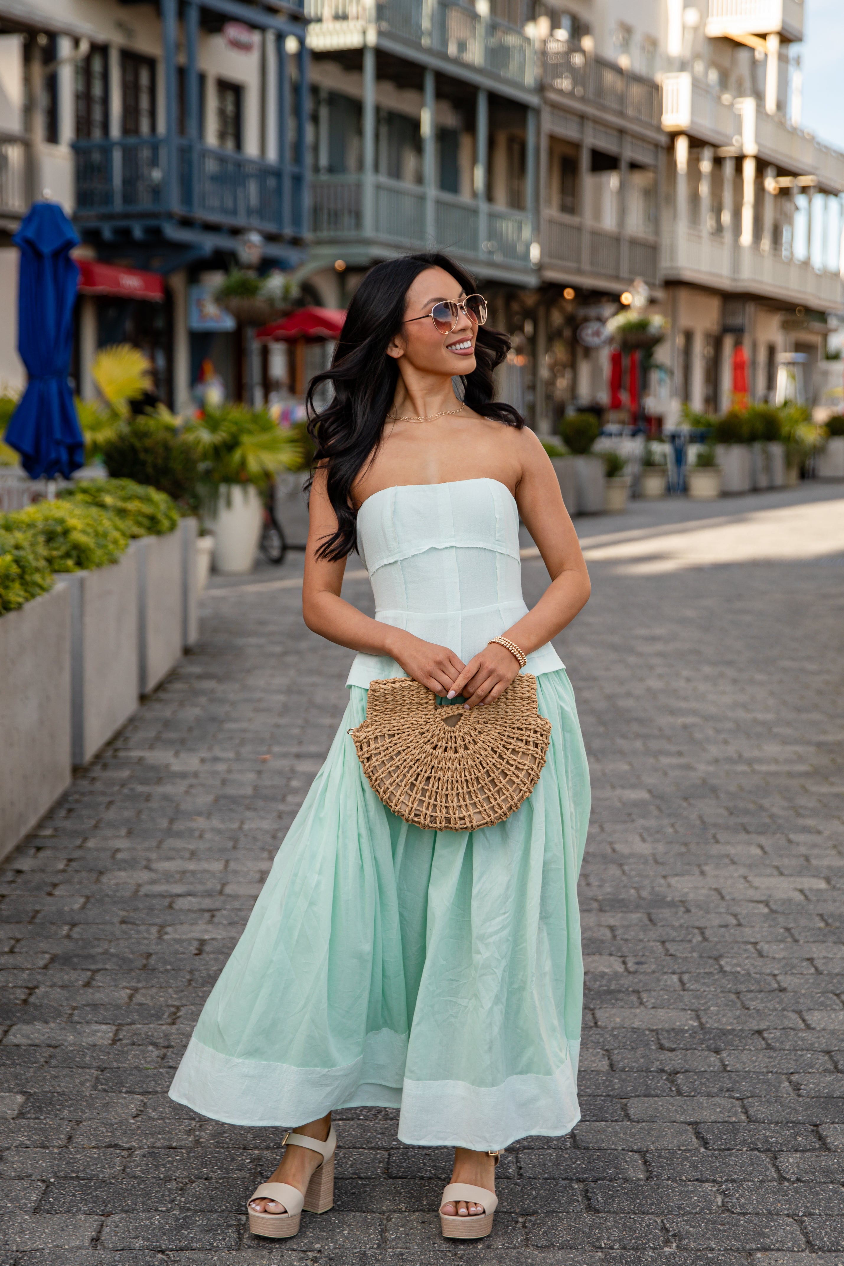 Woman in a strapless white top and light green skirt with a woven handbag on a street.