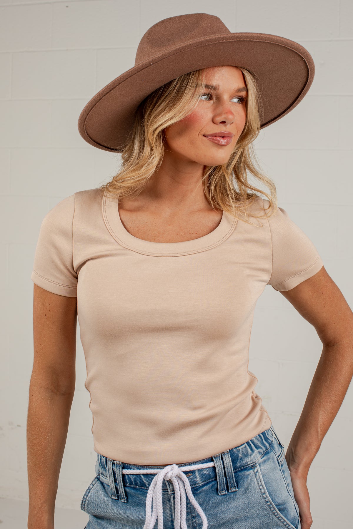 A woman wearing a beige short-sleeve scoop-neck t-shirt, blue jeans with a white drawstring, and a brown wide-brim hat, posing against a white paneled wall.