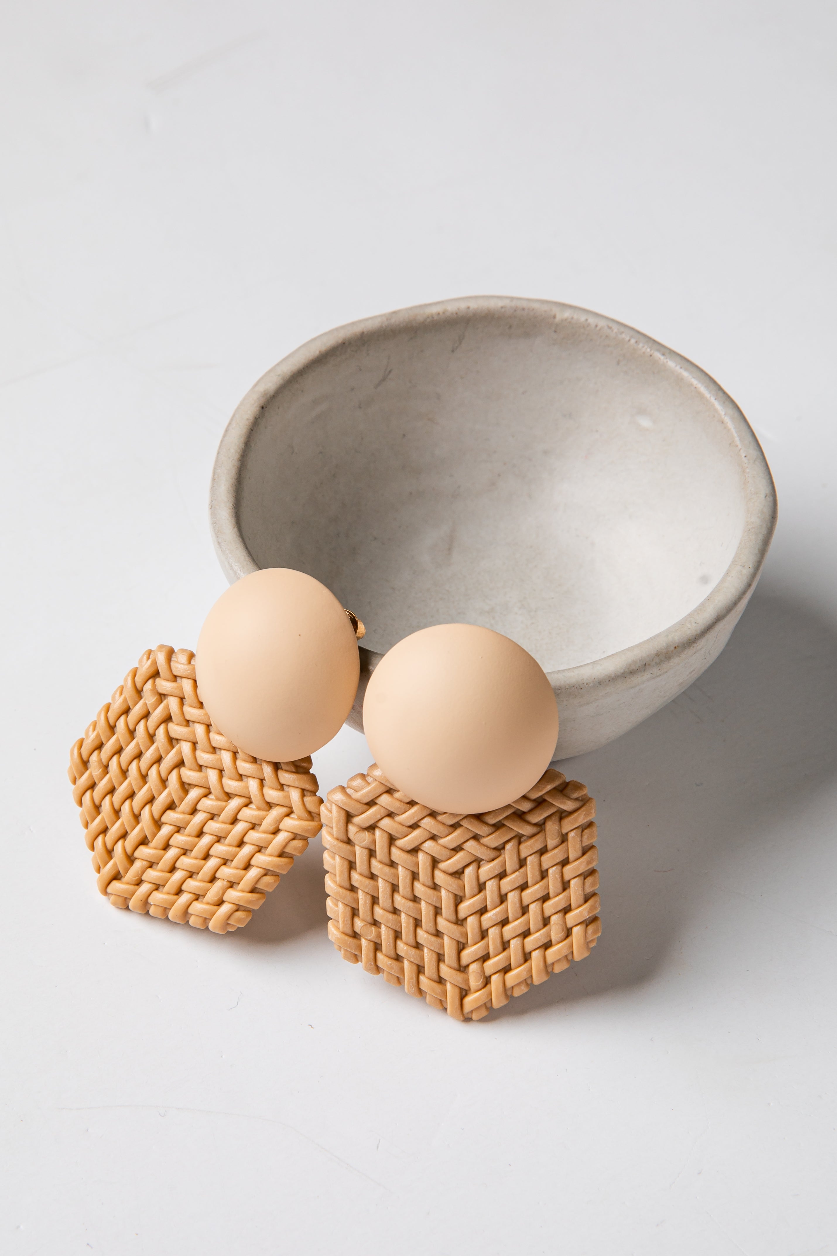 Pair of earrings with woven square and round beige beads in a ceramic bowl on a light background