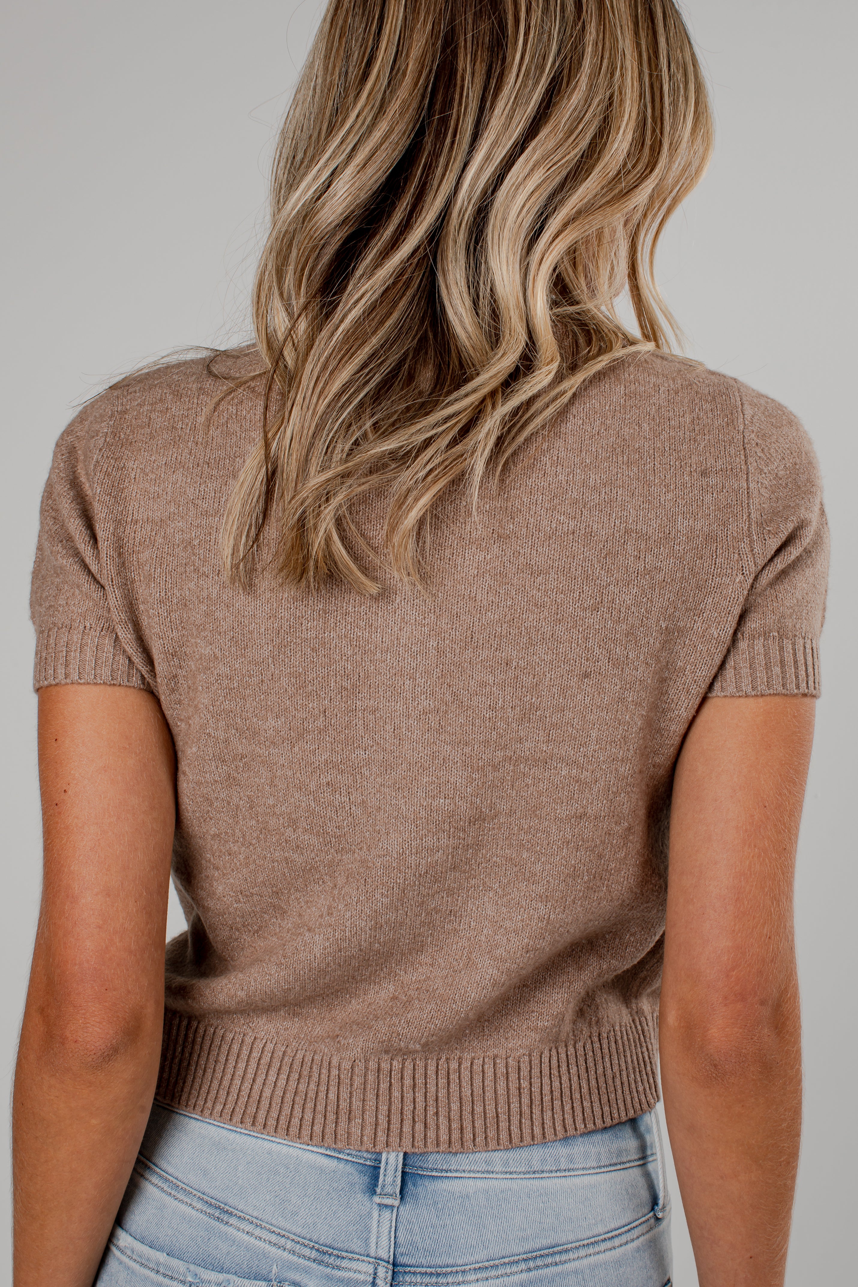 A woman viewed from the back wearing a short-sleeve knit top in a warm taupe-brown color, paired with light blue jeans, against a plain light gray studio background.