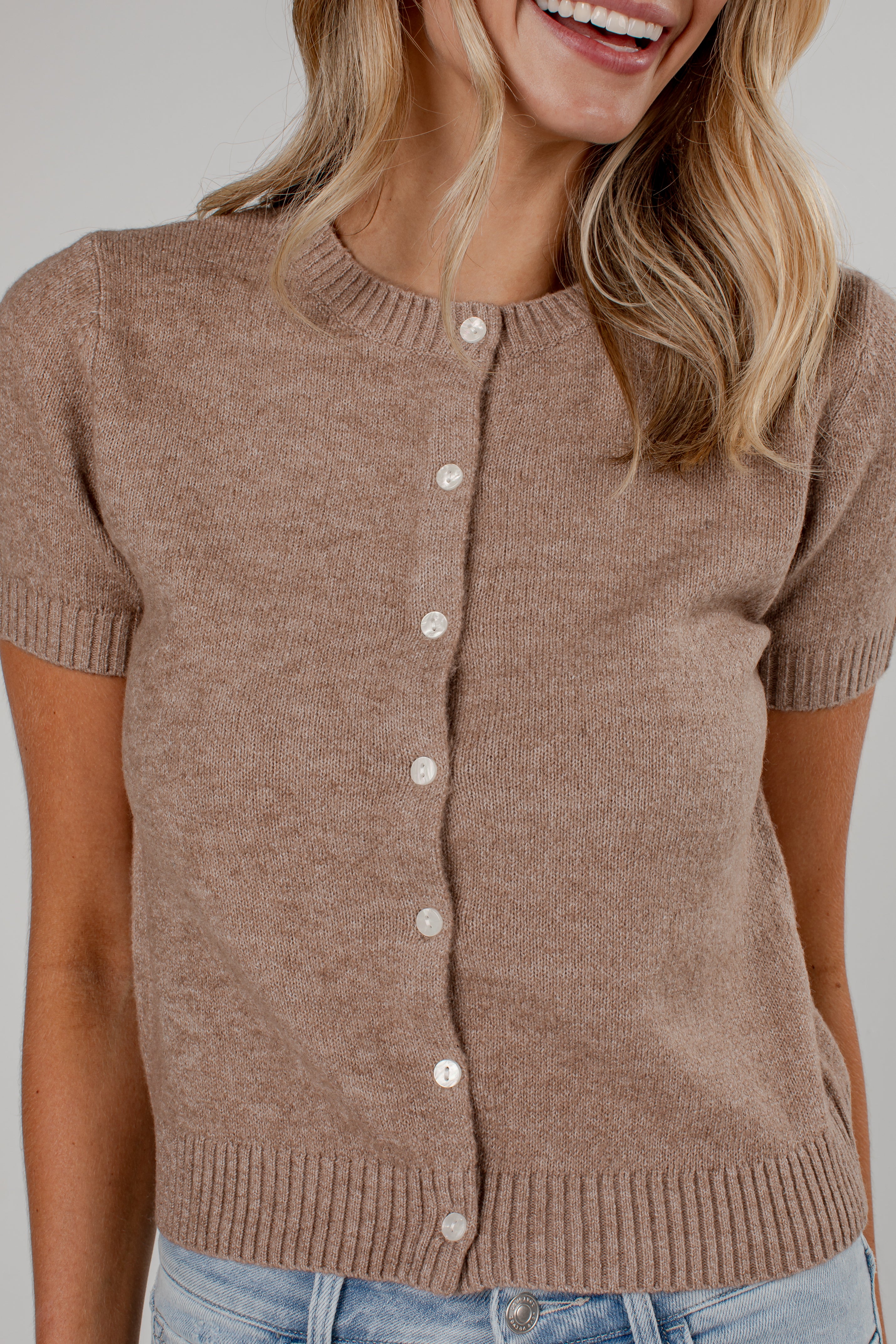 A woman wearing a short-sleeve knit cardigan in a warm taupe-brown color, featuring a button-up front with white buttons and ribbed trim, paired with light blue jeans.