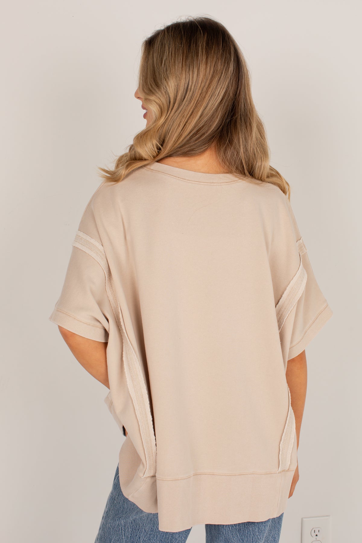 A woman viewed from behind wearing an oversized short-sleeve top in a light beige or nude color, paired with blue jeans, against a plain off-white wall.