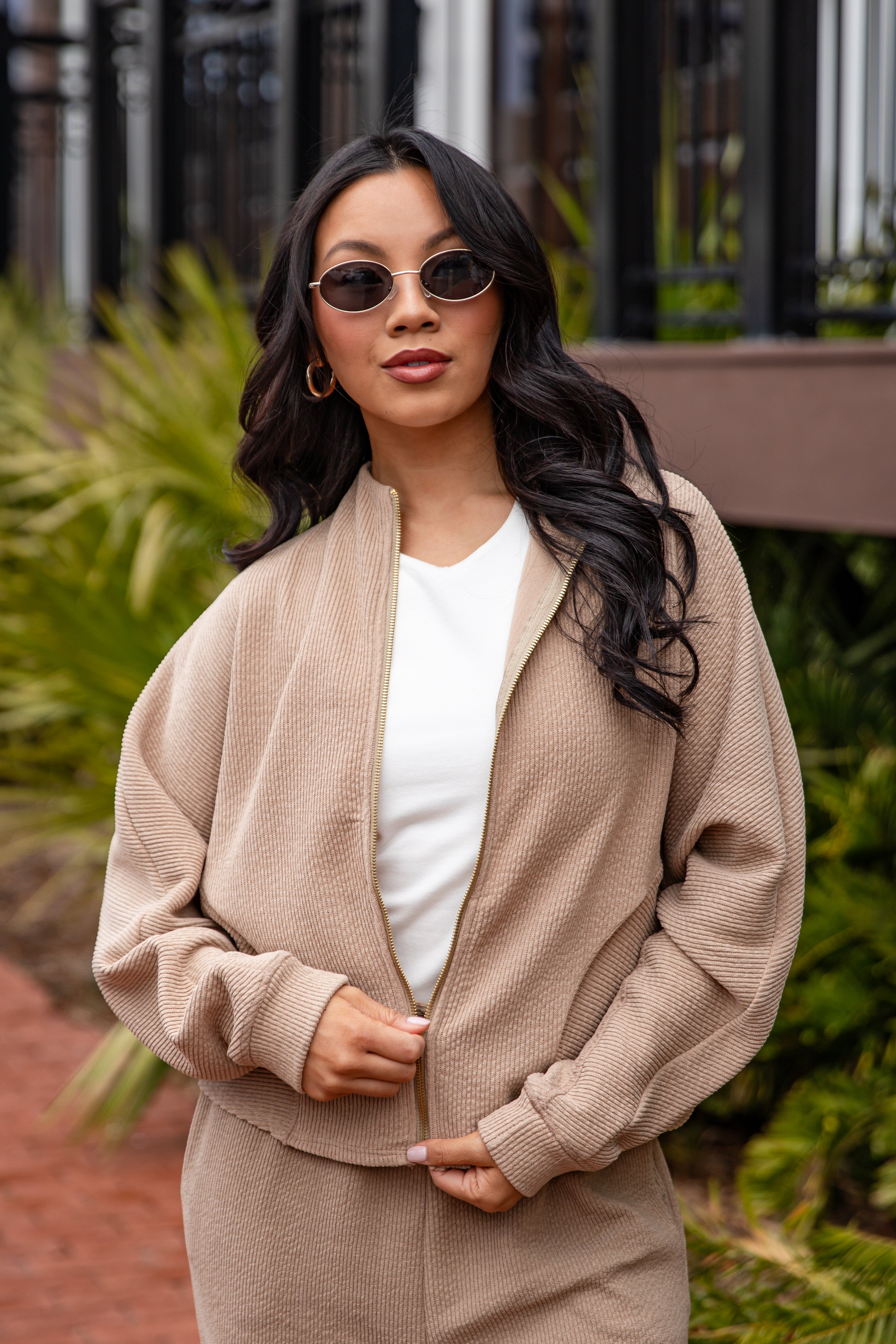 Woman wearing a beige cardigan over a white top, standing outdoors with greenery in the background.