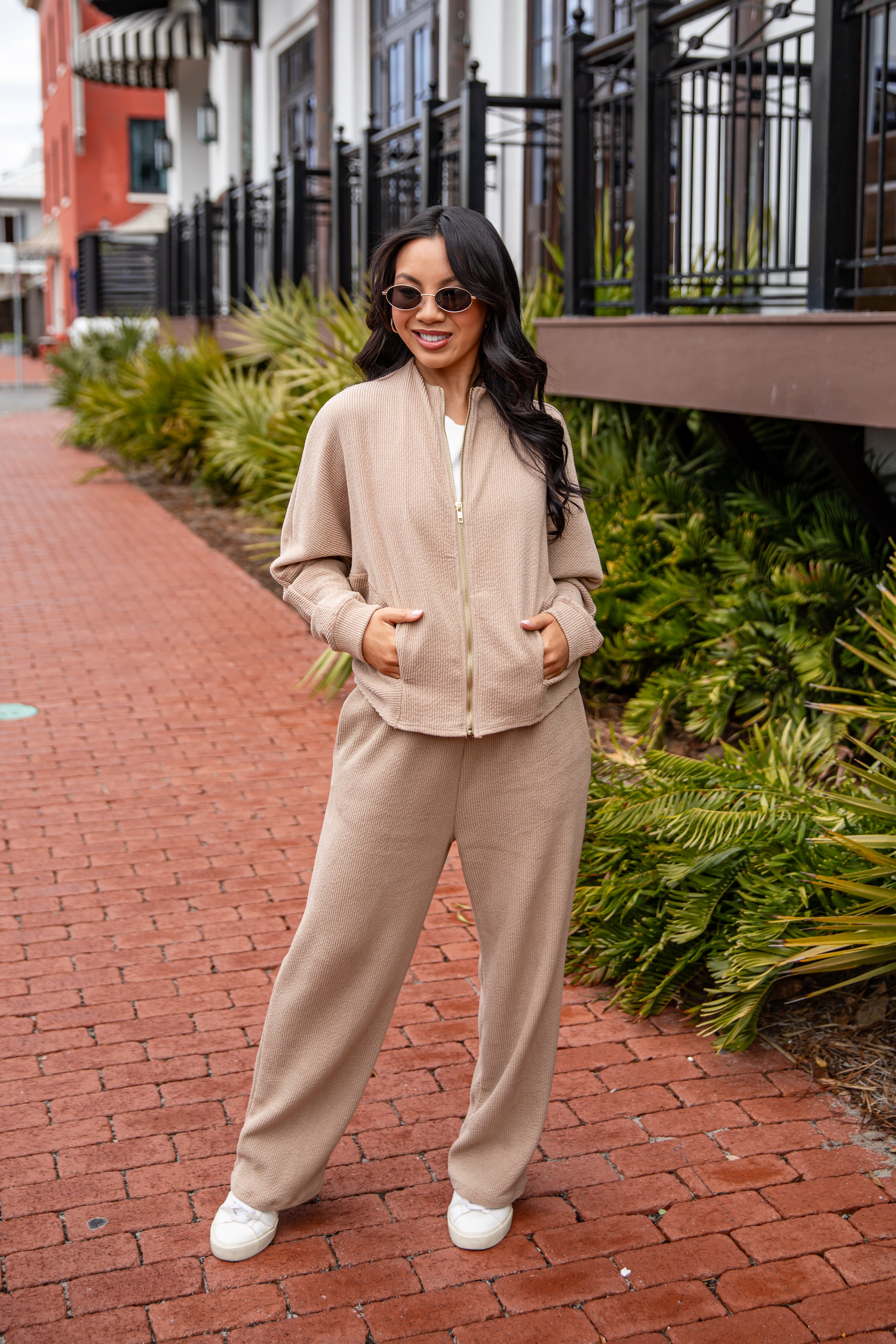 Woman in a beige outfit standing on a brick sidewalk with greenery and buildings in the background