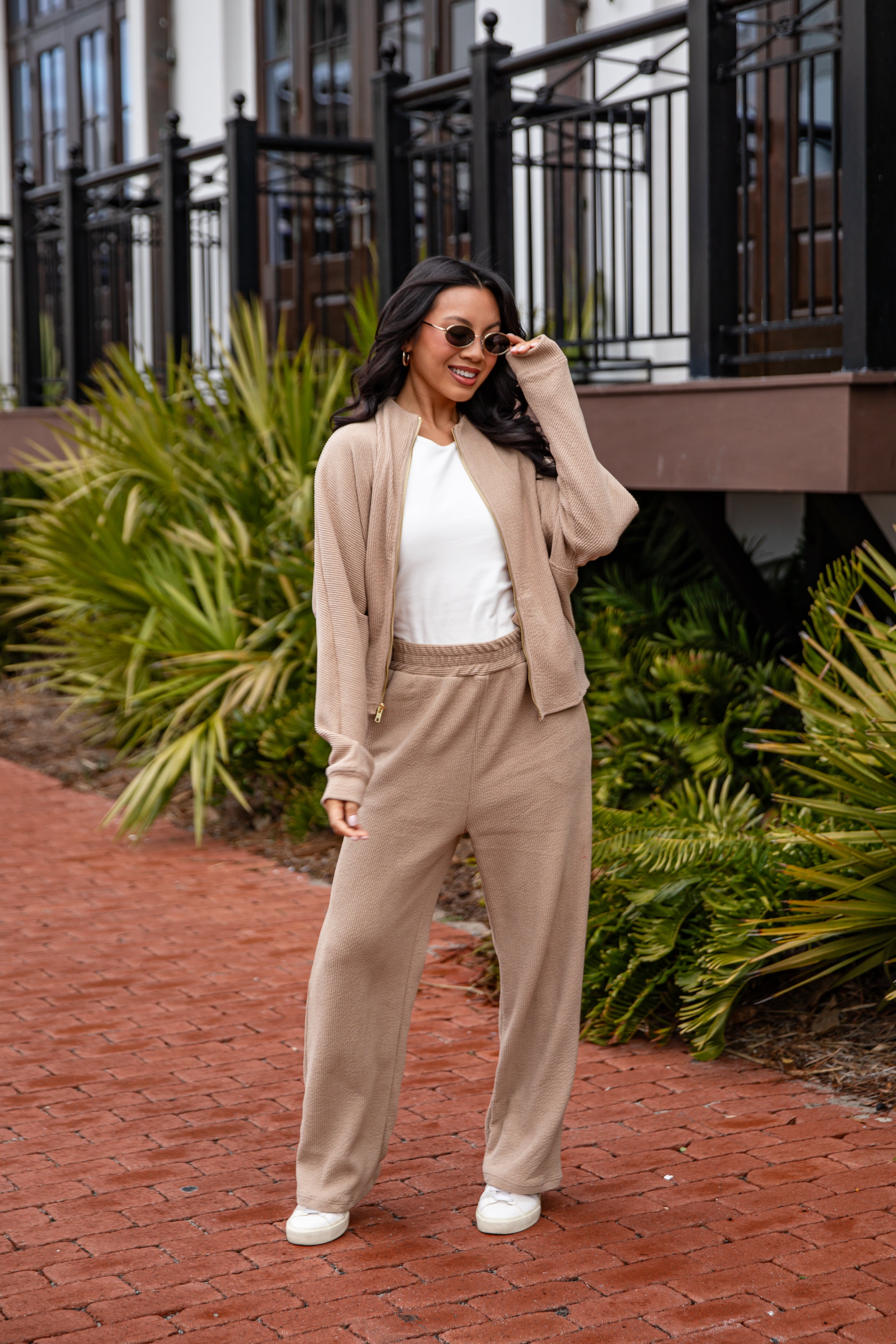 Woman in beige cardigan and pants standing on a brick path with greenery in the background