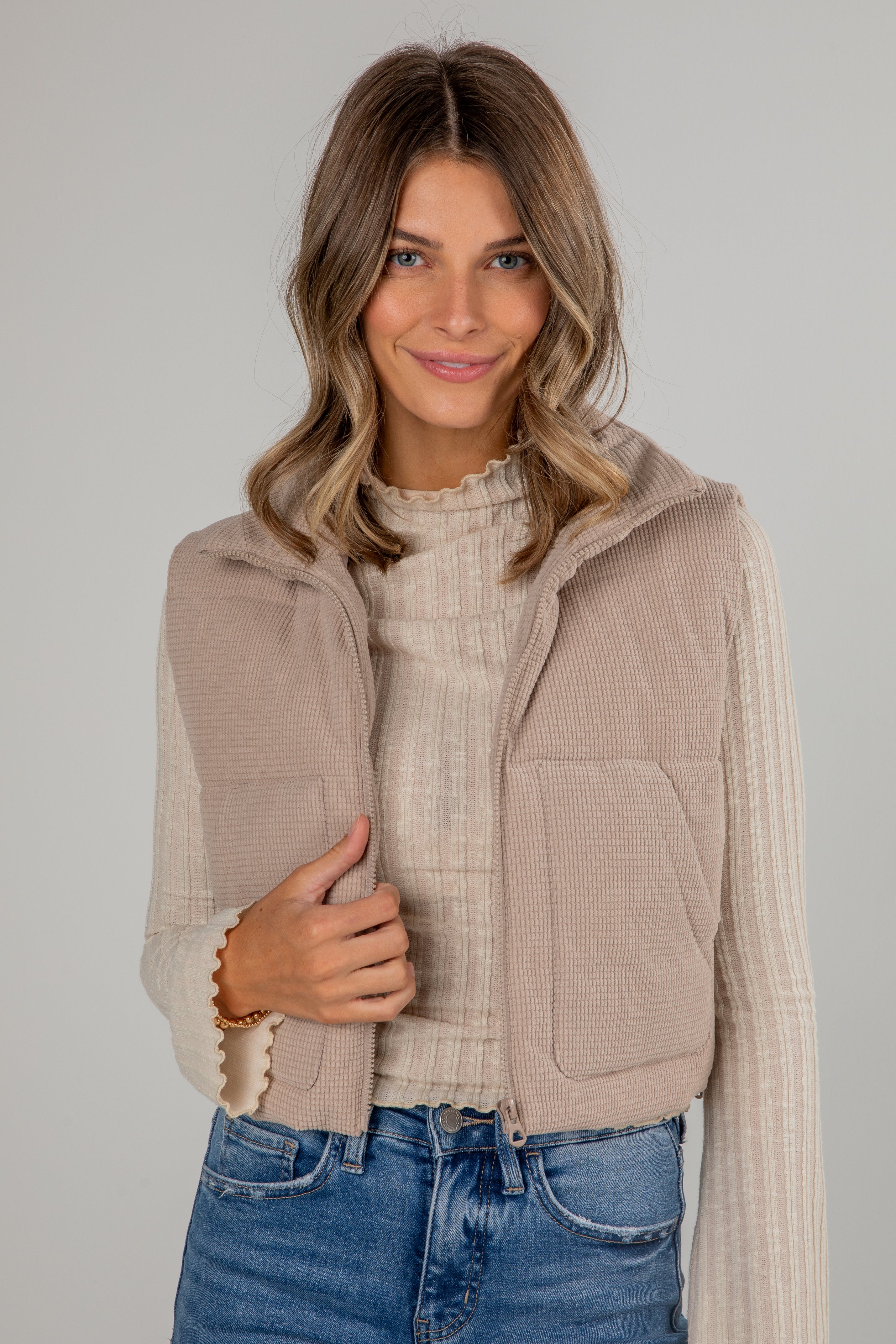 Woman wearing a beige vest over a sweater and jeans on a plain background