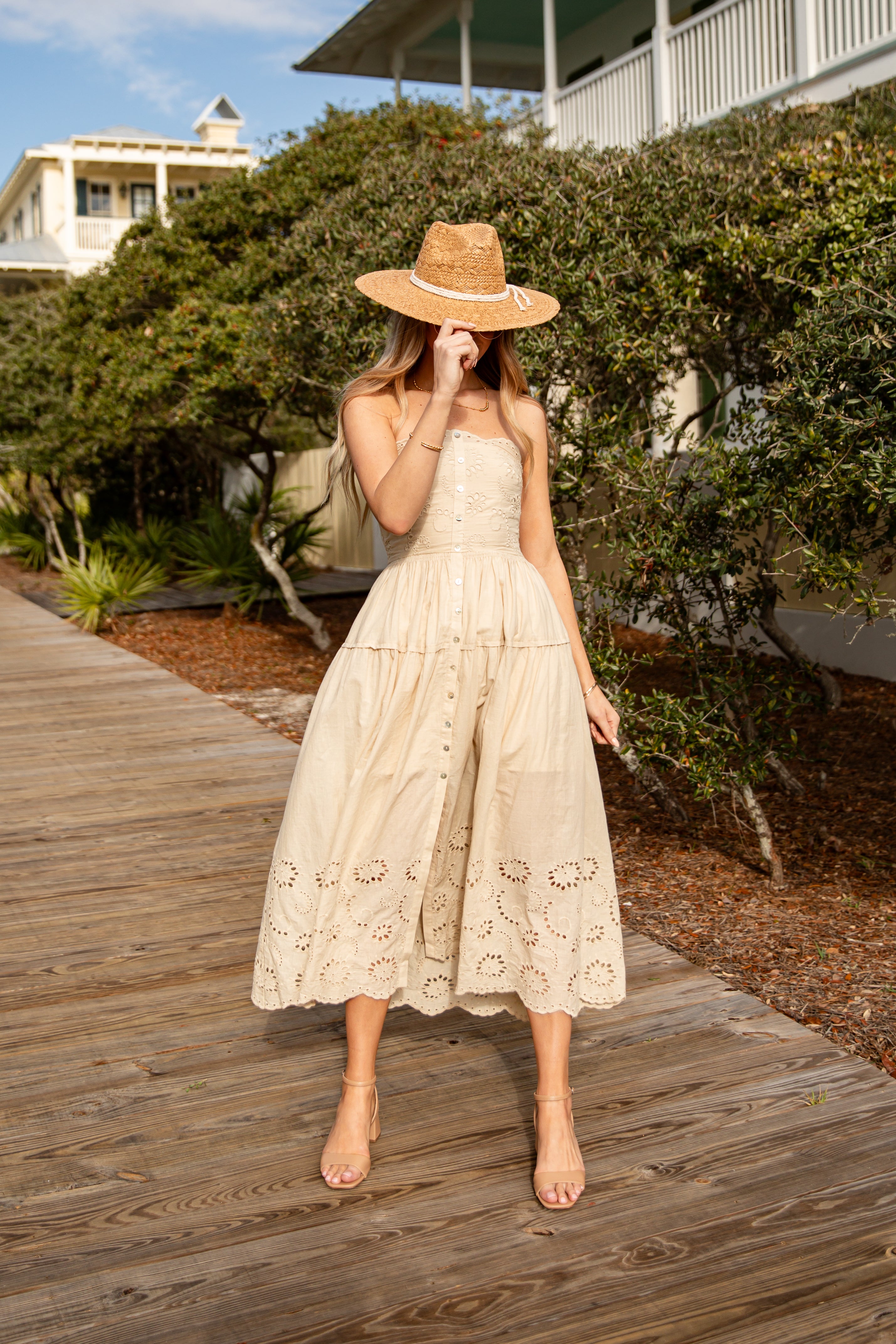 Woman in a beige lace dress and straw hat standing on a wooden boardwalk.