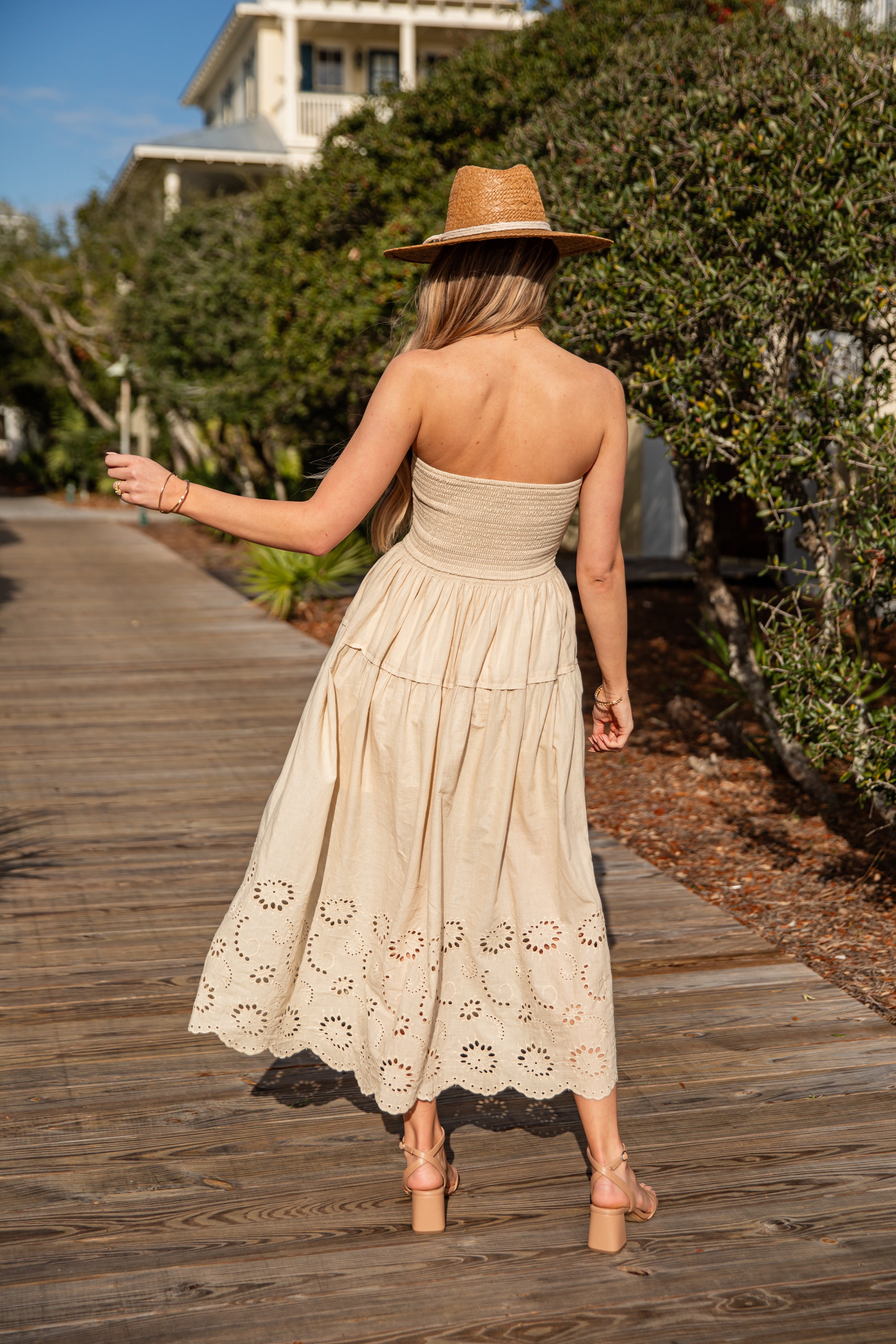 Woman in a beige strapless dress and hat walking on a wooden boardwalk.