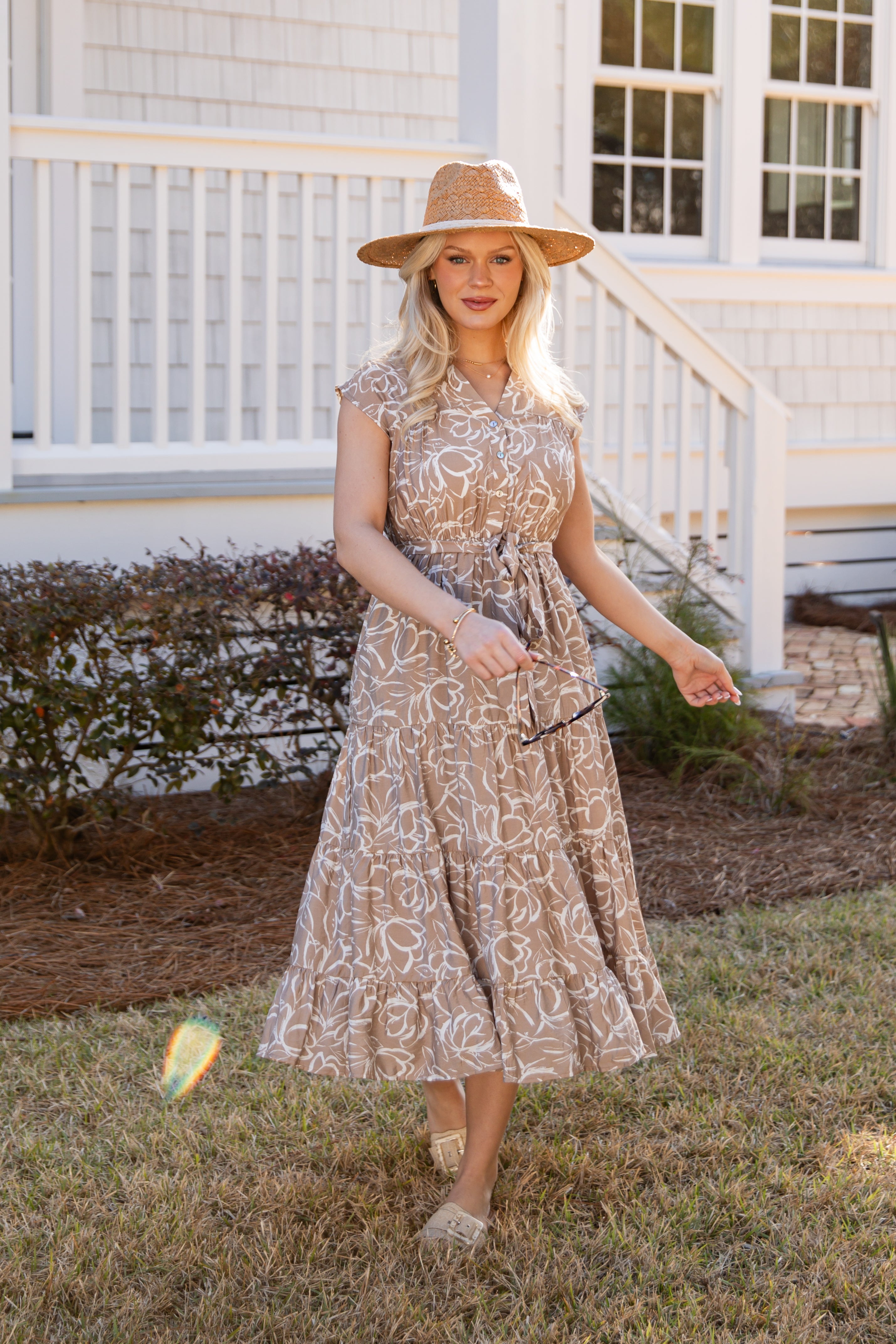 Woman in a patterned dress and hat standing in front of a house.