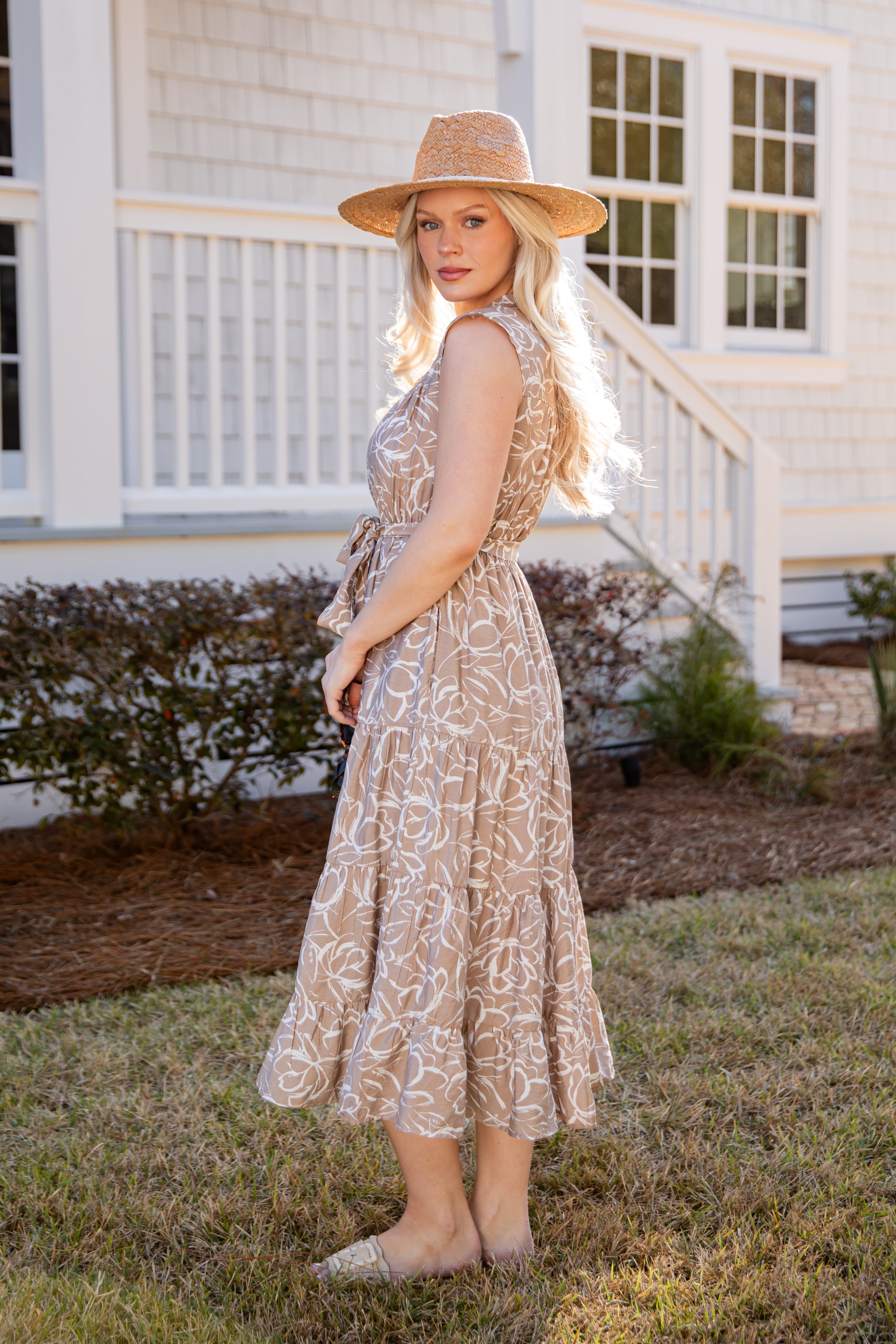 Woman in a patterned dress and hat standing in front of a house.