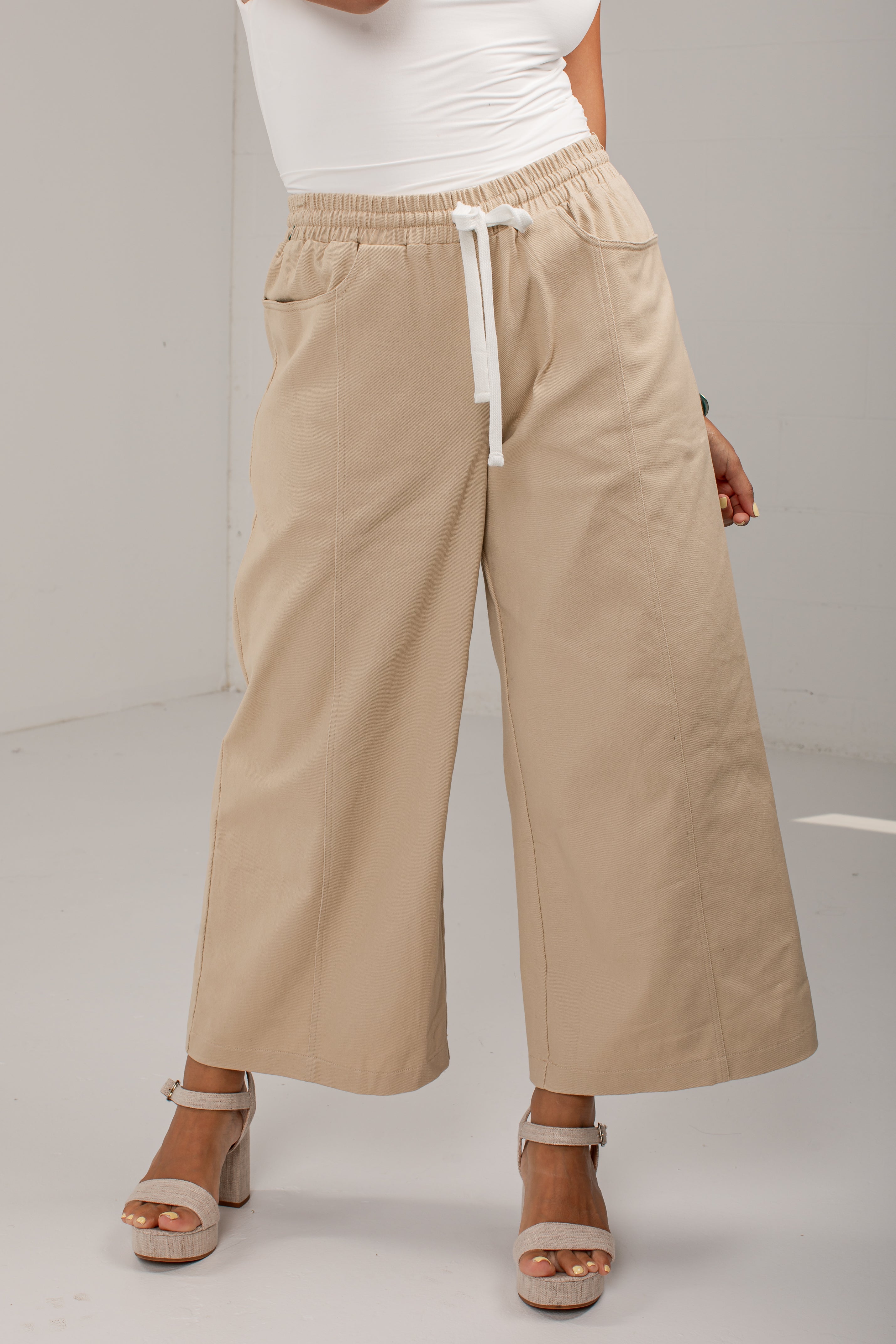 A person wearing beige wide-leg cropped pants with an elastic waistband and white drawstring, paired with a white top and beige heeled sandals, photographed in a studio setting.