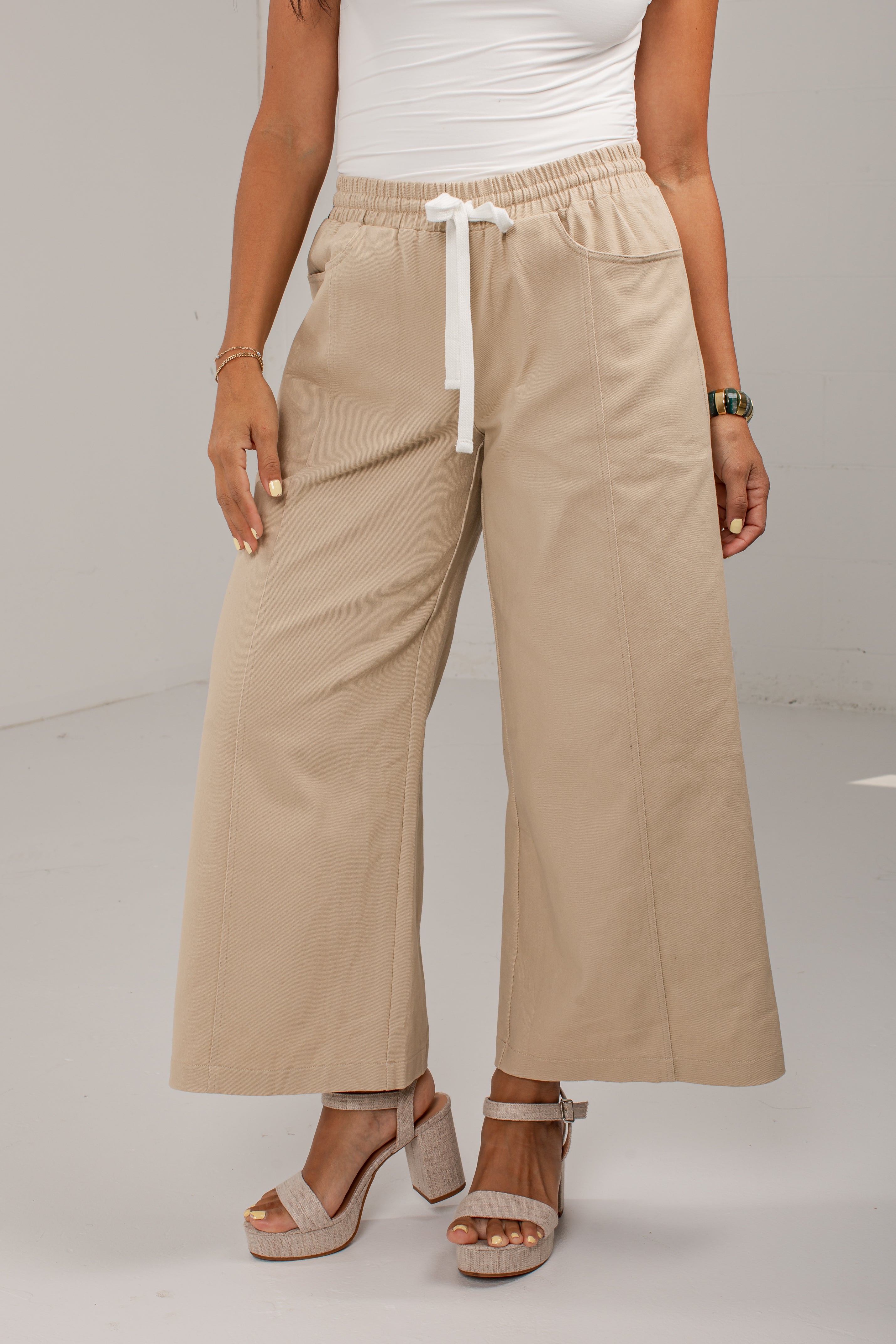 A model wearing beige wide-leg cropped pants with an elastic waistband and white drawstring, paired with a white top and beige platform sandals.