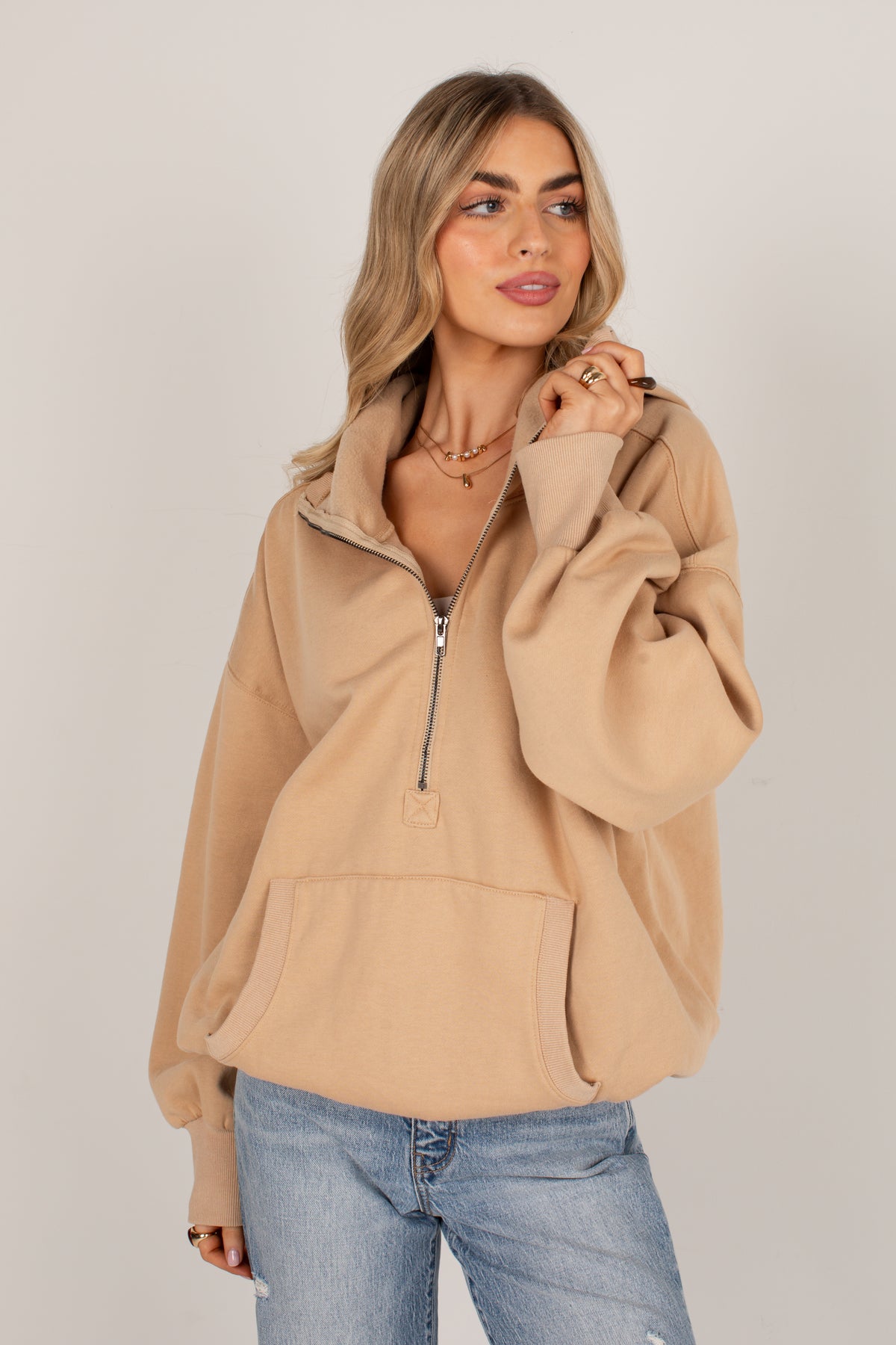 A woman wearing a beige half-zip hoodie with a high collar and front pockets, paired with light-wash blue jeans, photographed against a plain light gray studio background.
