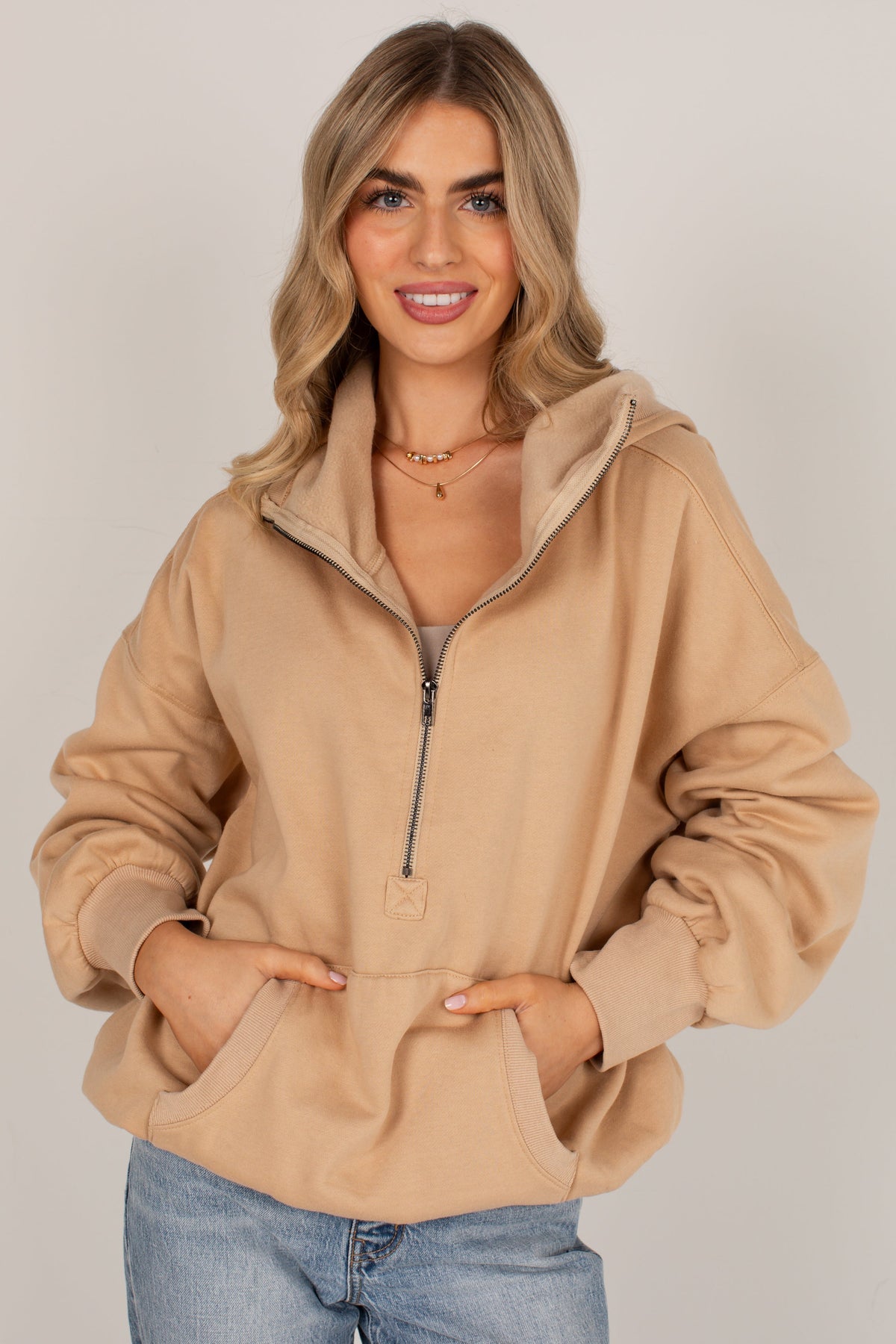 A woman modeling a beige half-zip hoodie with a drawstring hood and front pockets, paired with light blue jeans, against a plain light gray studio background.