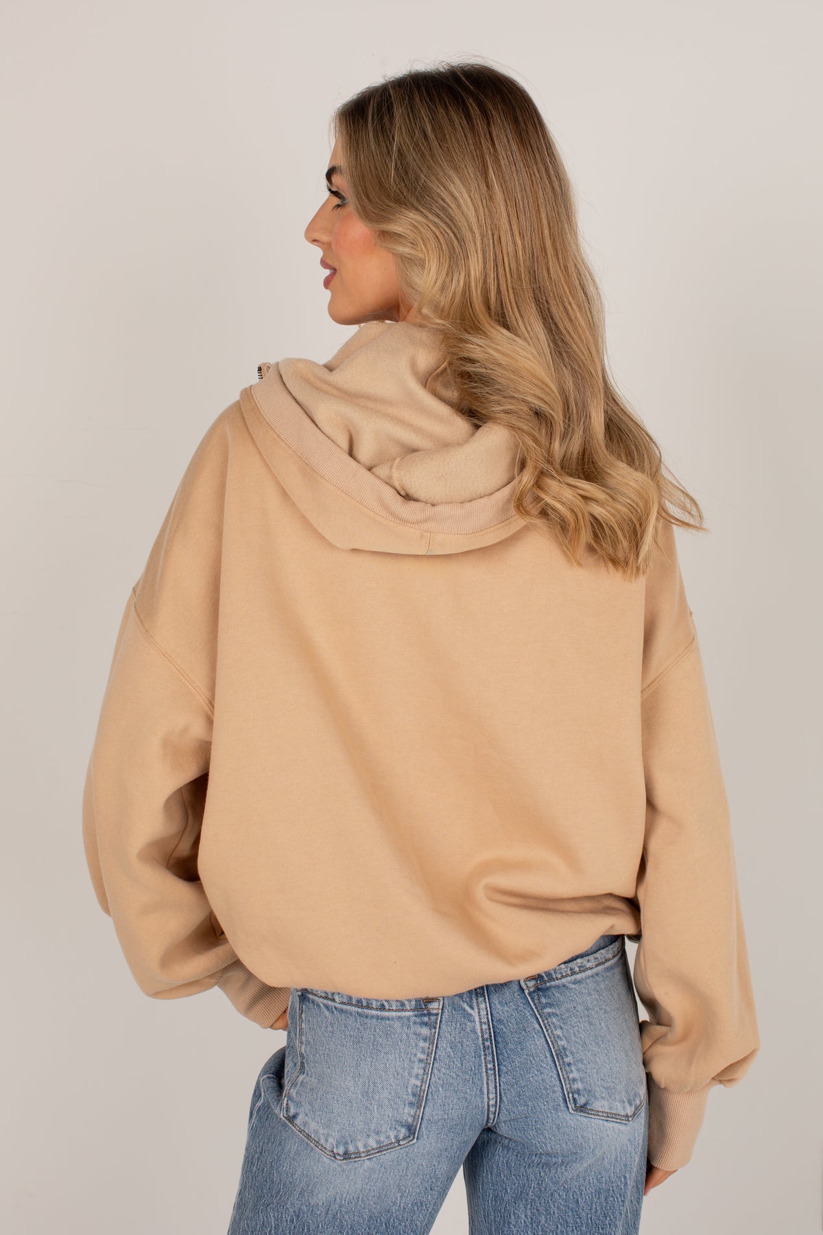 A woman shown from the back wearing a light tan oversized hoodie and blue jeans, photographed against a plain light gray studio background.