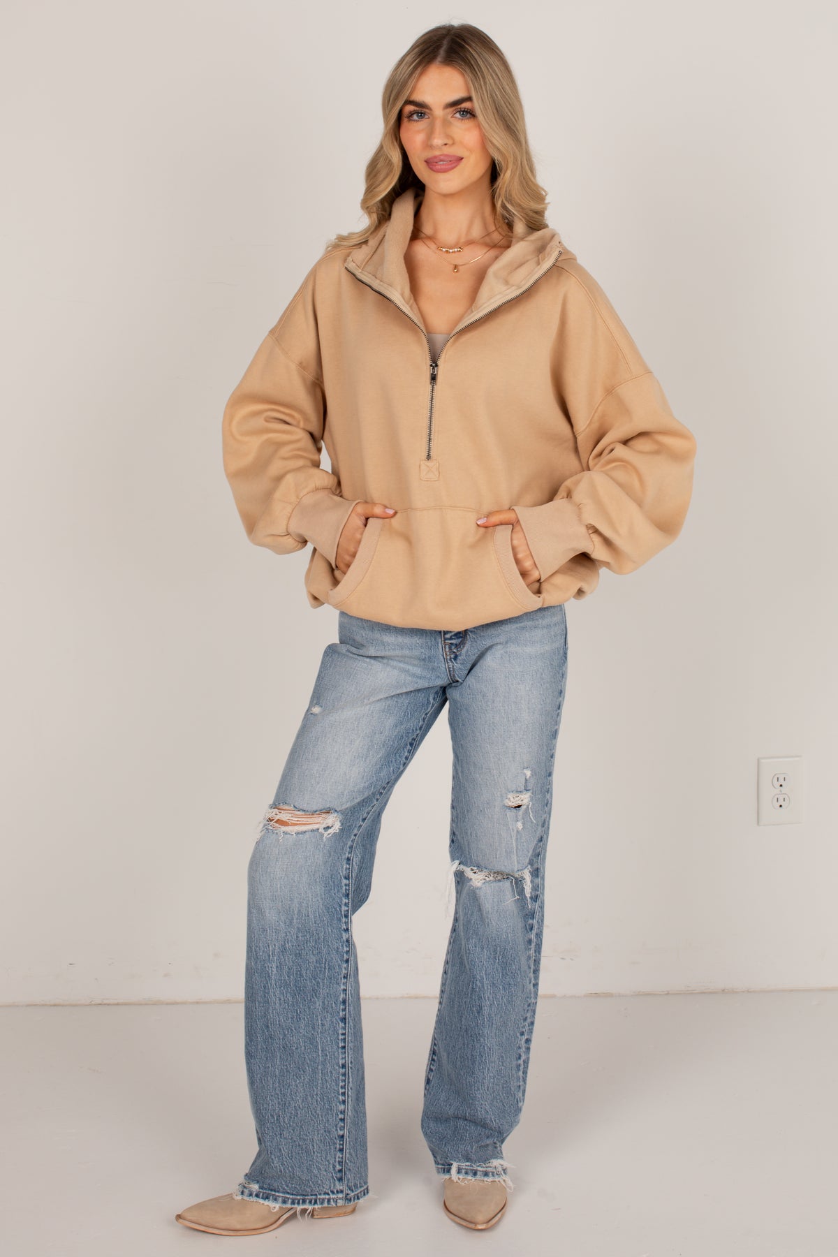 A woman modeling a beige half-zip fleece pullover with a high collar and kangaroo pockets, paired with distressed light-wash blue jeans and beige flats, standing against a plain off-white wall.