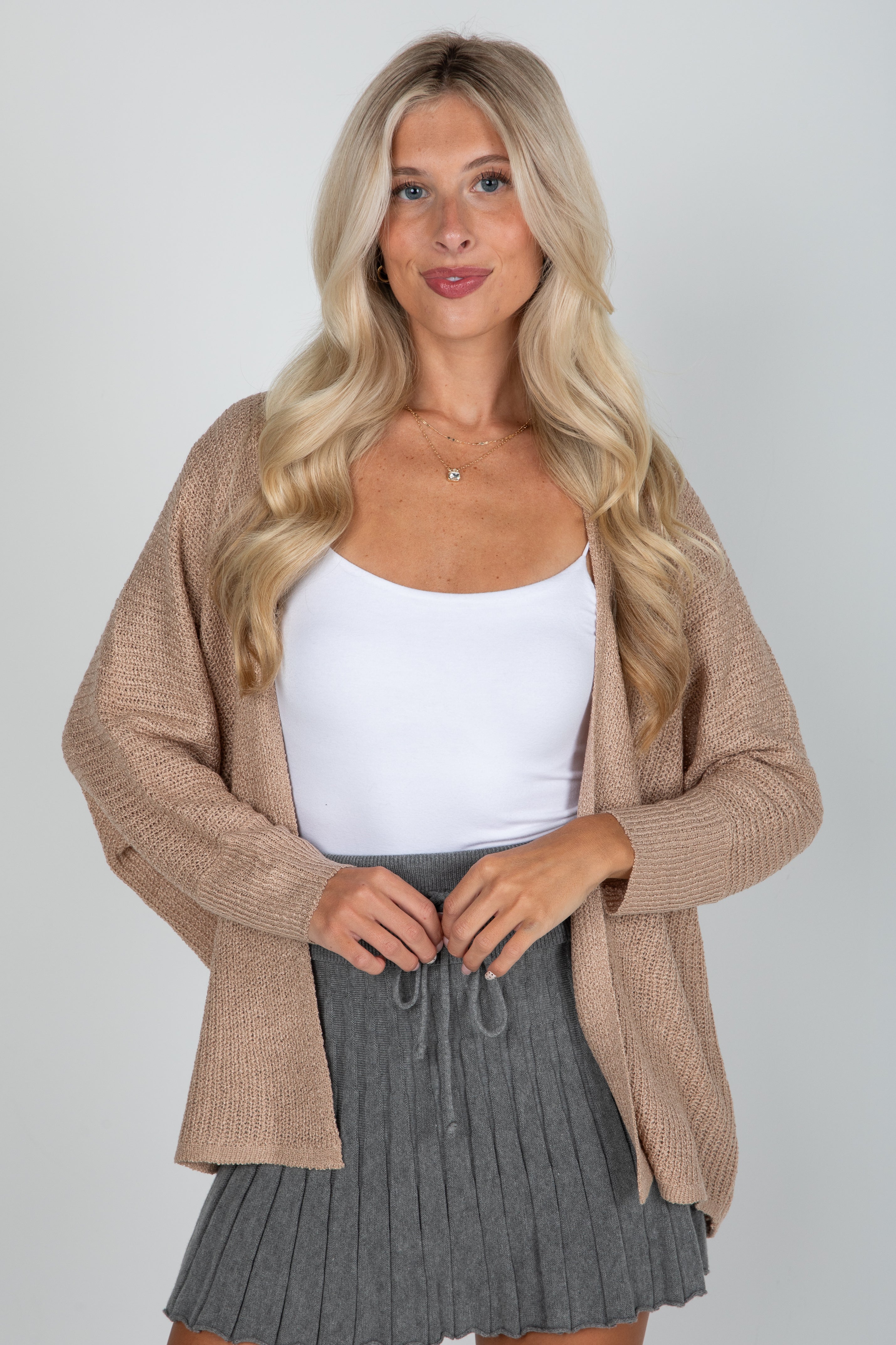 Woman wearing a beige cardigan, white top, and gray skirt on a plain background