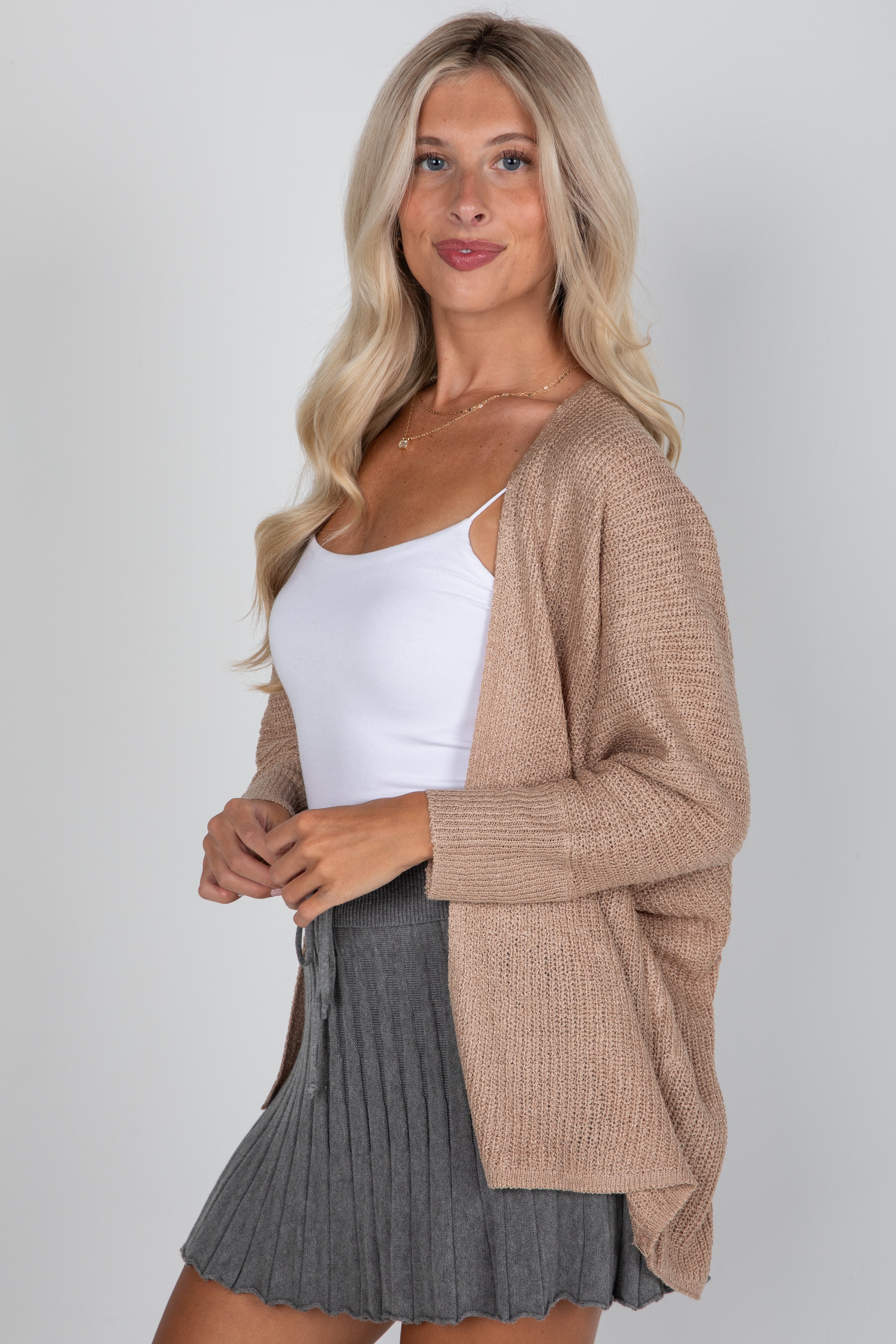 Woman wearing a beige cardigan over a white top and gray skirt on a plain background