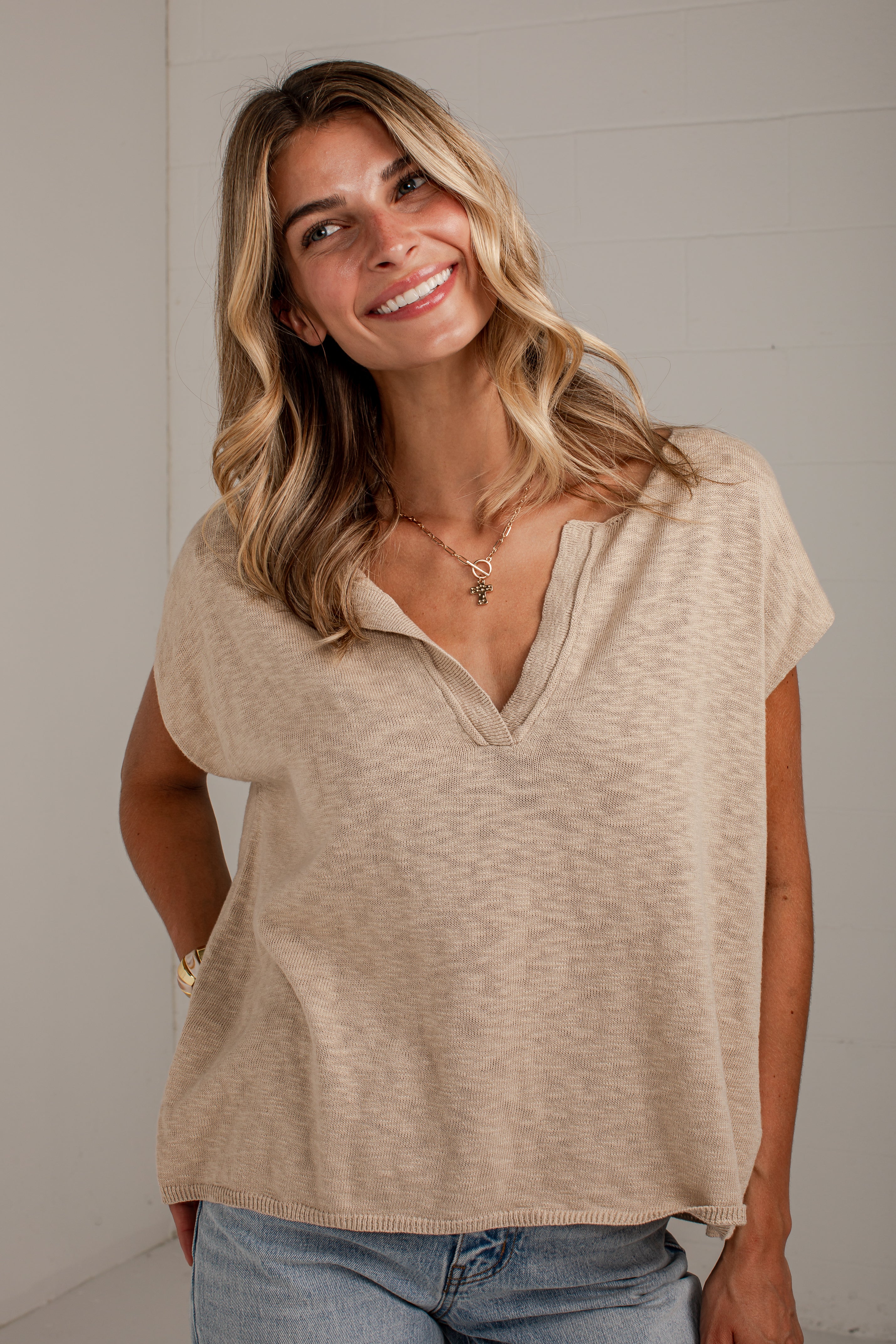 A woman with blonde hair wearing a beige short-sleeve knit top with a split V-neck and blue jeans, standing against a light gray wall.