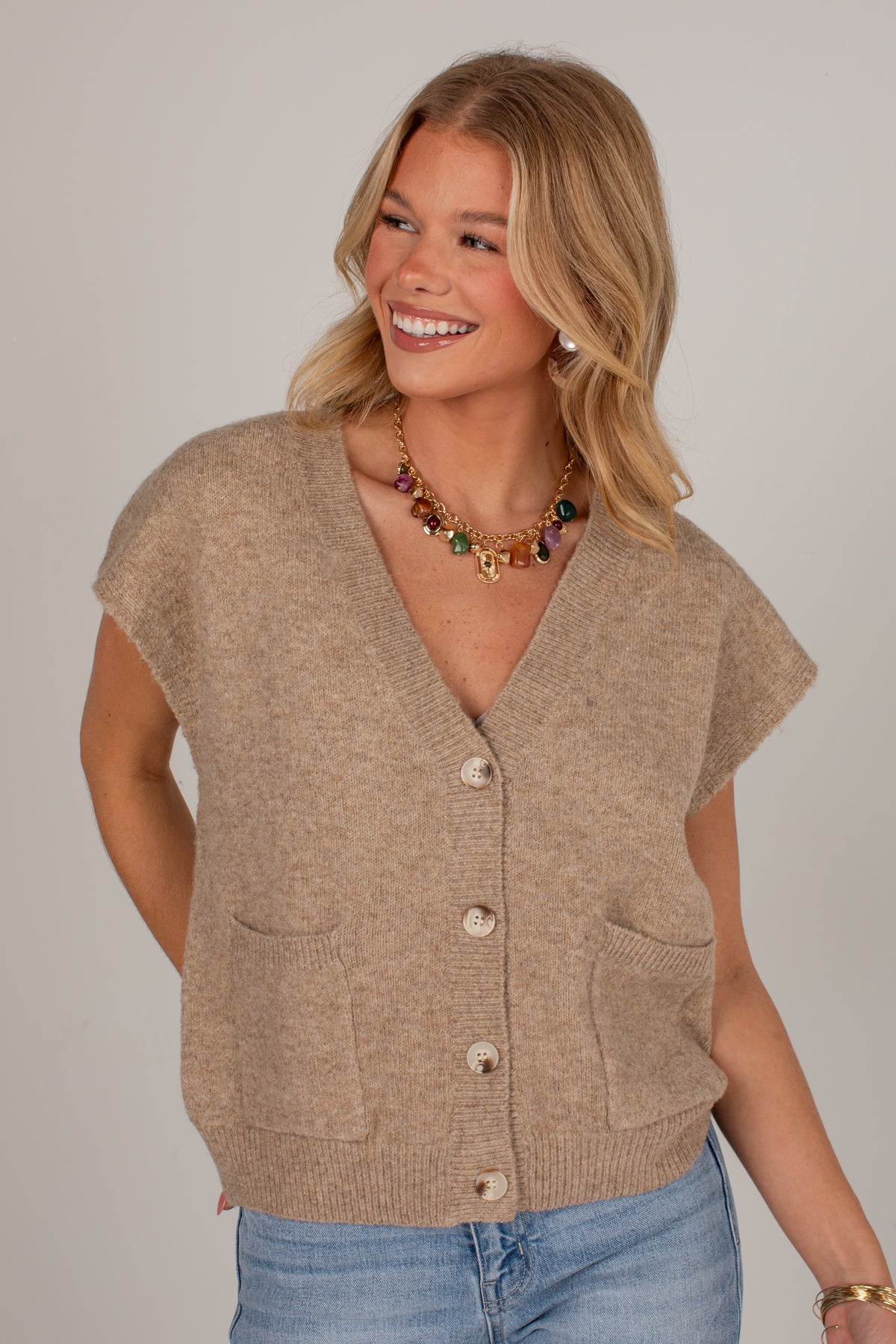 A smiling blonde woman wearing a beige knit cardigan vest with short sleeves, front buttons, and patch pockets, paired with light blue jeans against a plain light gray studio background.