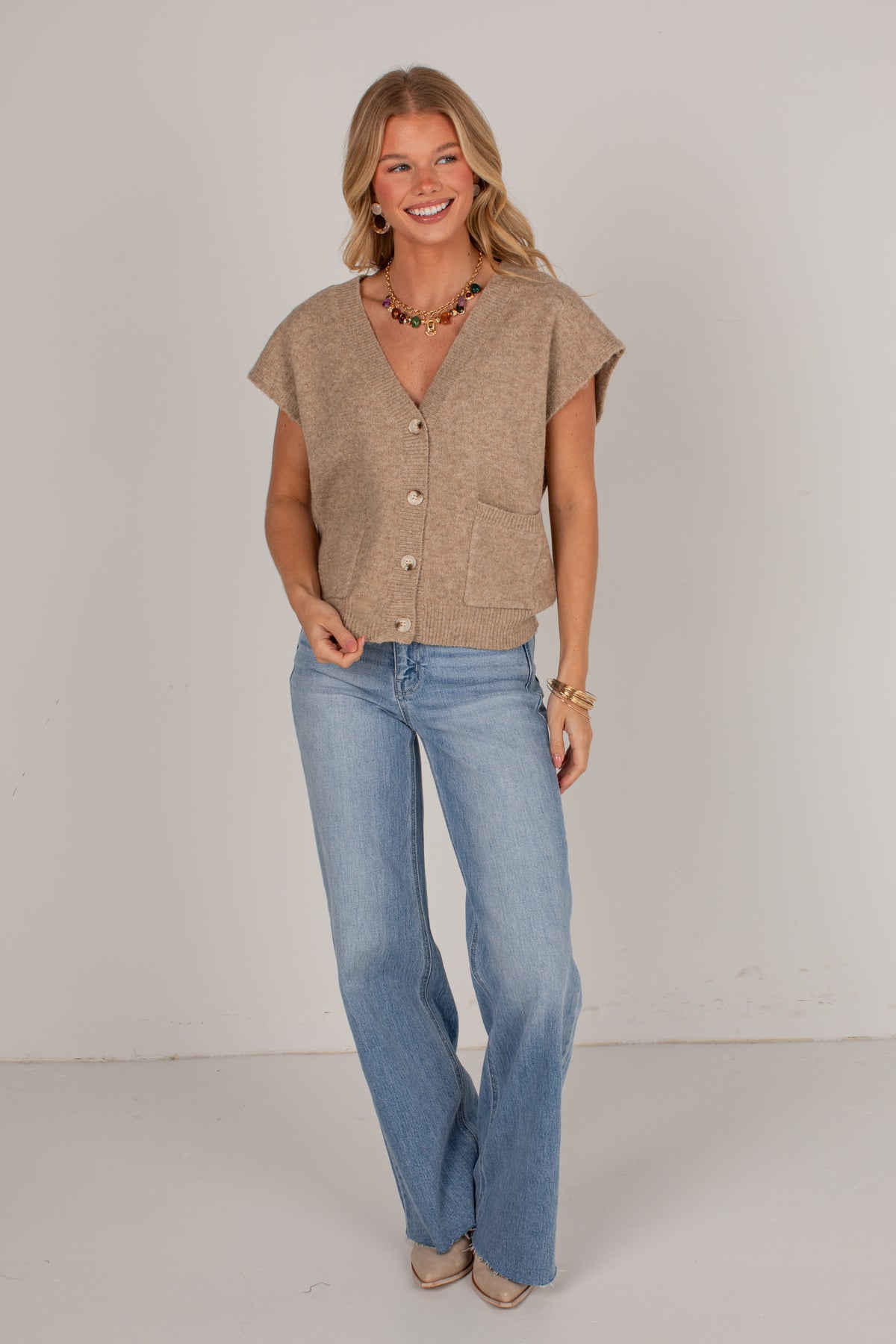 A woman modeling a beige short-sleeve button-front knit top with front pockets, paired with light-wash blue jeans, standing against a plain light gray studio background.