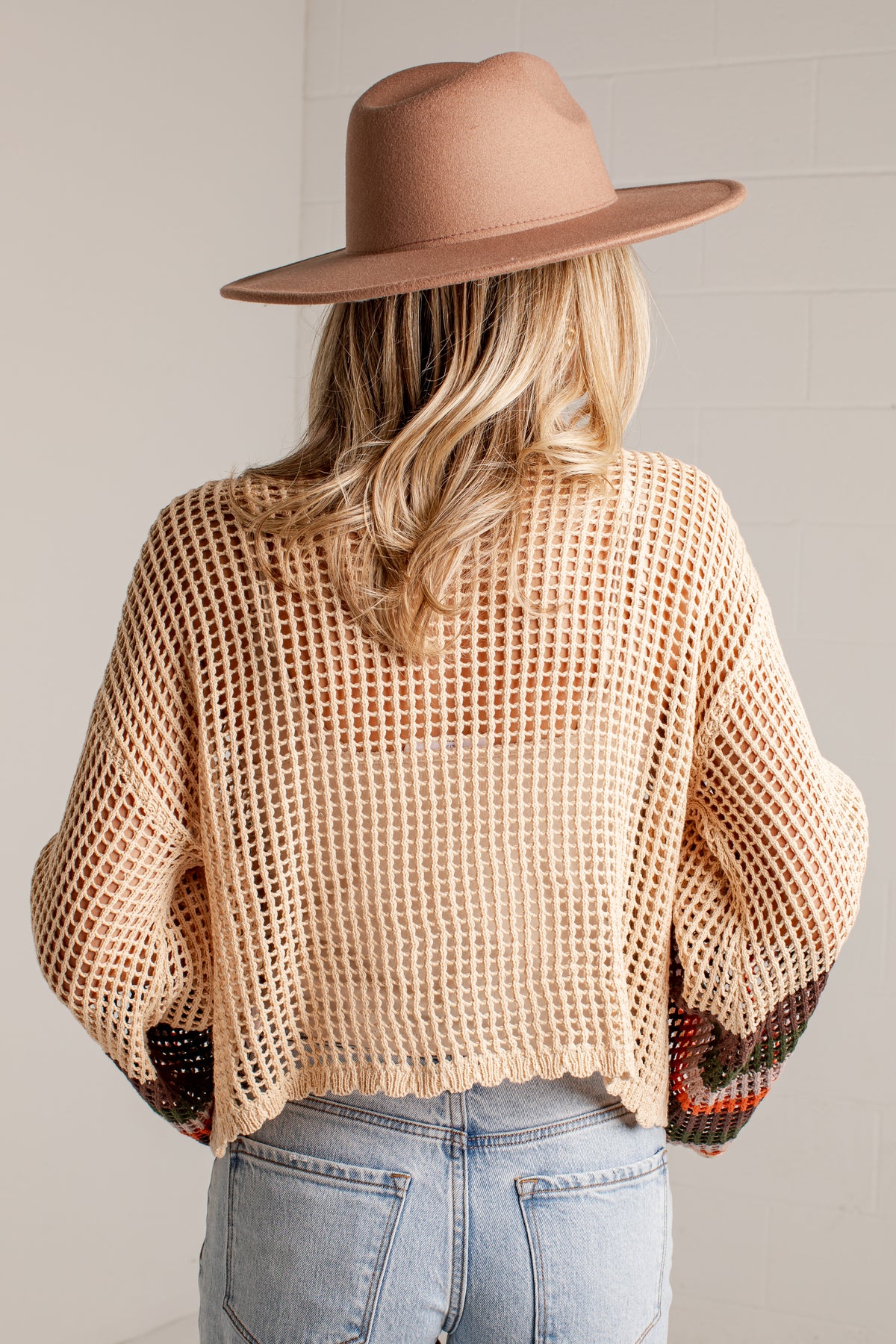 A woman shown from behind wearing a beige open-knit cropped sweater, a tan wide-brim hat, and light-wash blue jeans against a light gray wall.