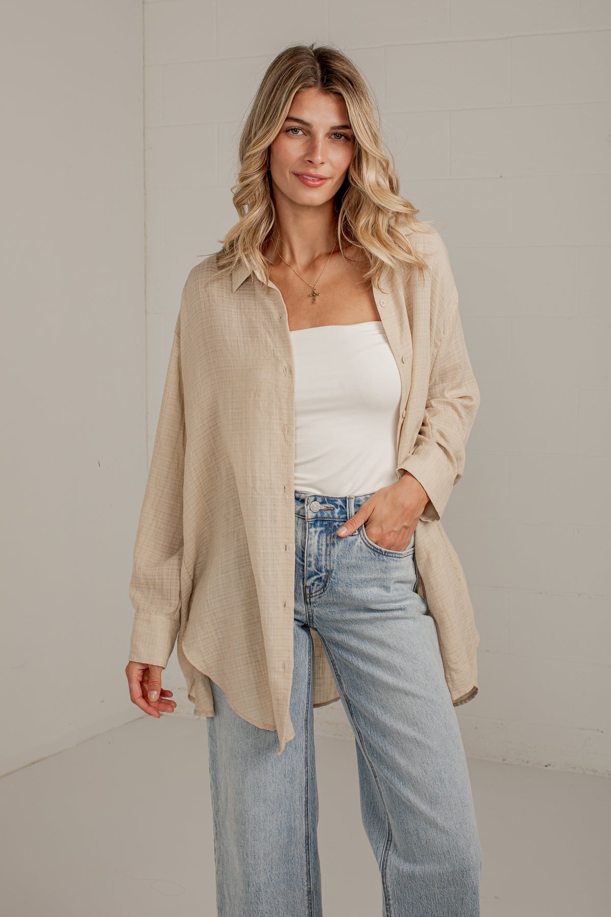 A woman modeling a light beige linen button-up shirt over a white tank top and light-wash blue jeans, standing against a neutral off-white wall.