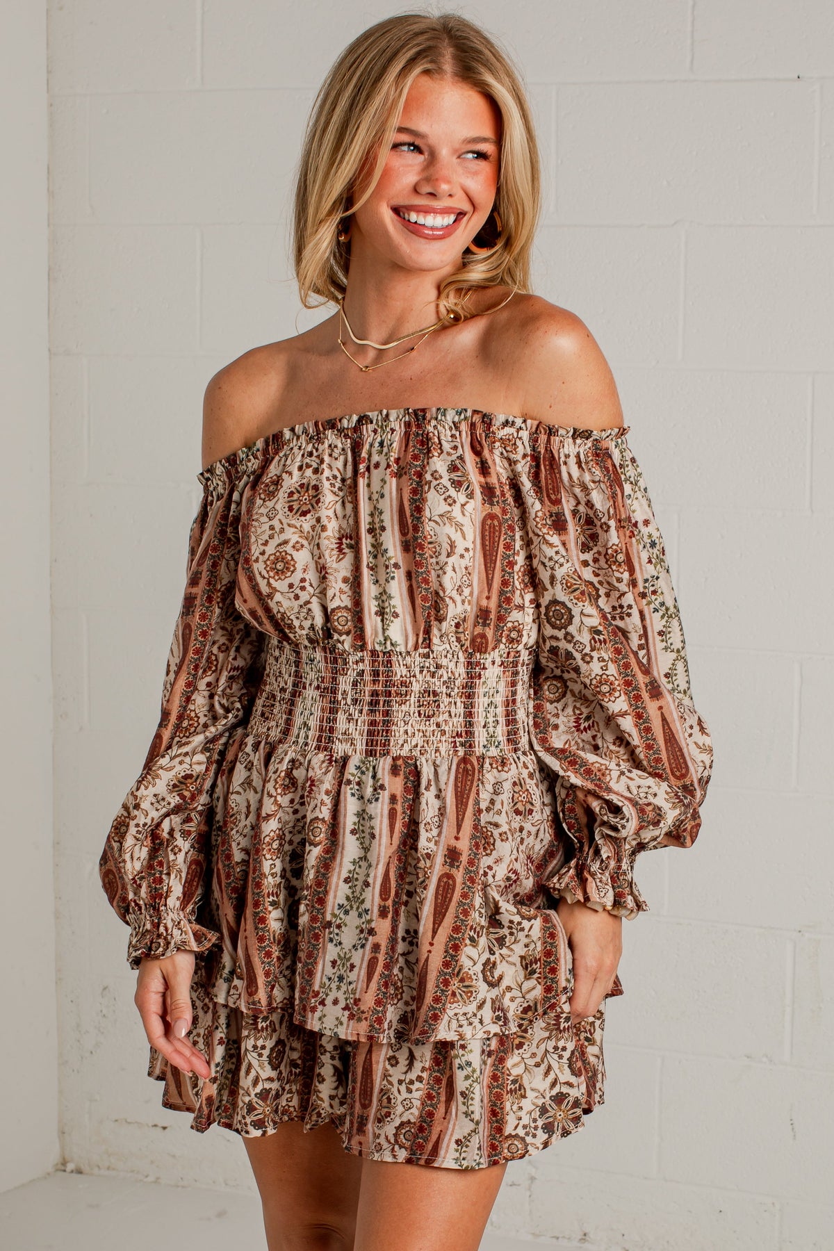 A woman modeling an off-the-shoulder mini dress with a brown and beige paisley pattern, paired with beige ankle boots, standing against a white brick wall.