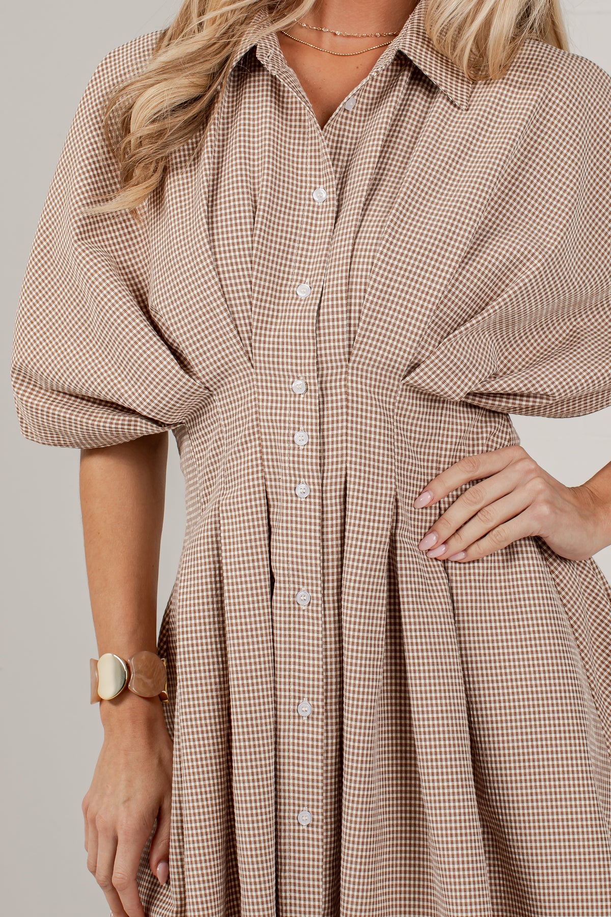 A close-up of a woman wearing a beige and brown gingham button-up shirt dress with voluminous elbow-length sleeves and a pleated skirt, photographed against a plain light gray studio background.