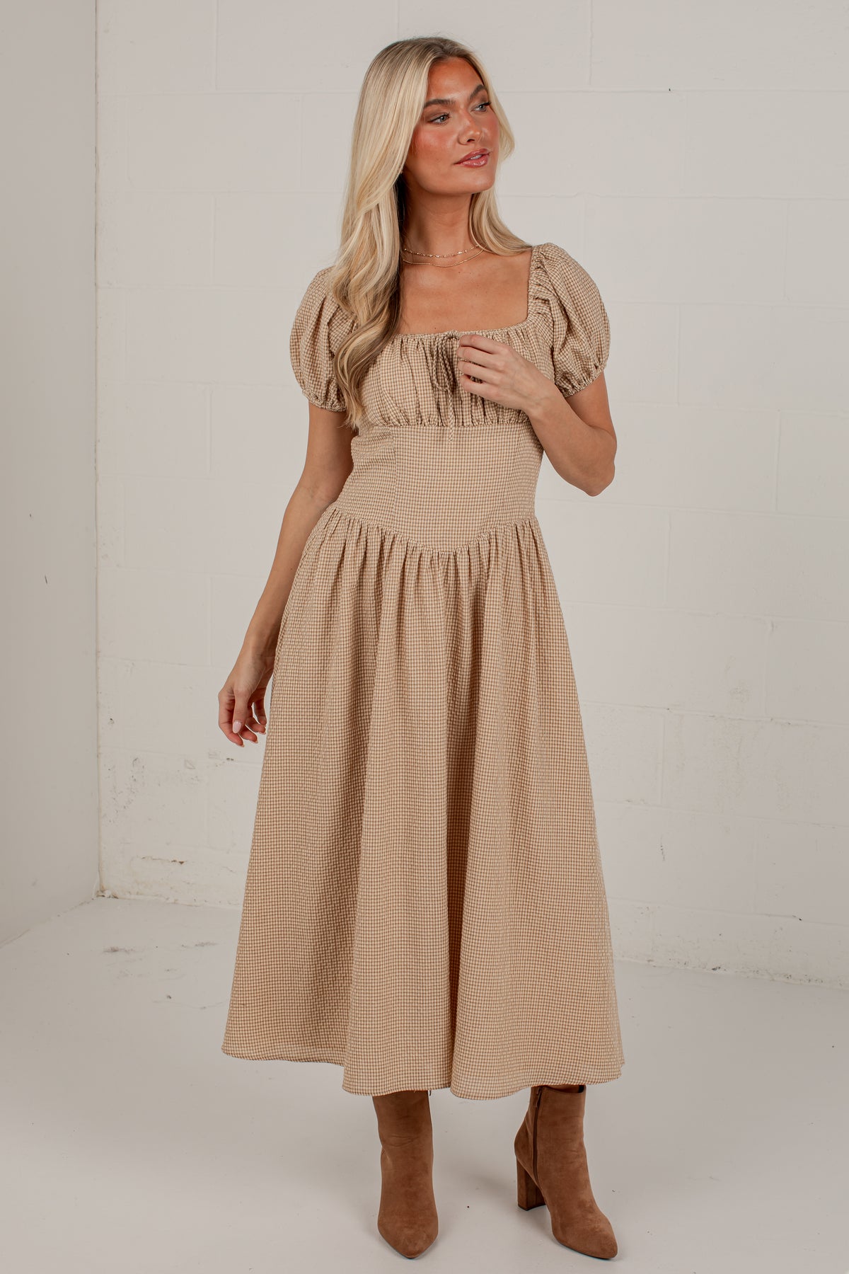 A woman modeling a beige midi dress with puff sleeves, a square neckline, and a gathered skirt, paired with brown ankle boots in a bright studio setting.