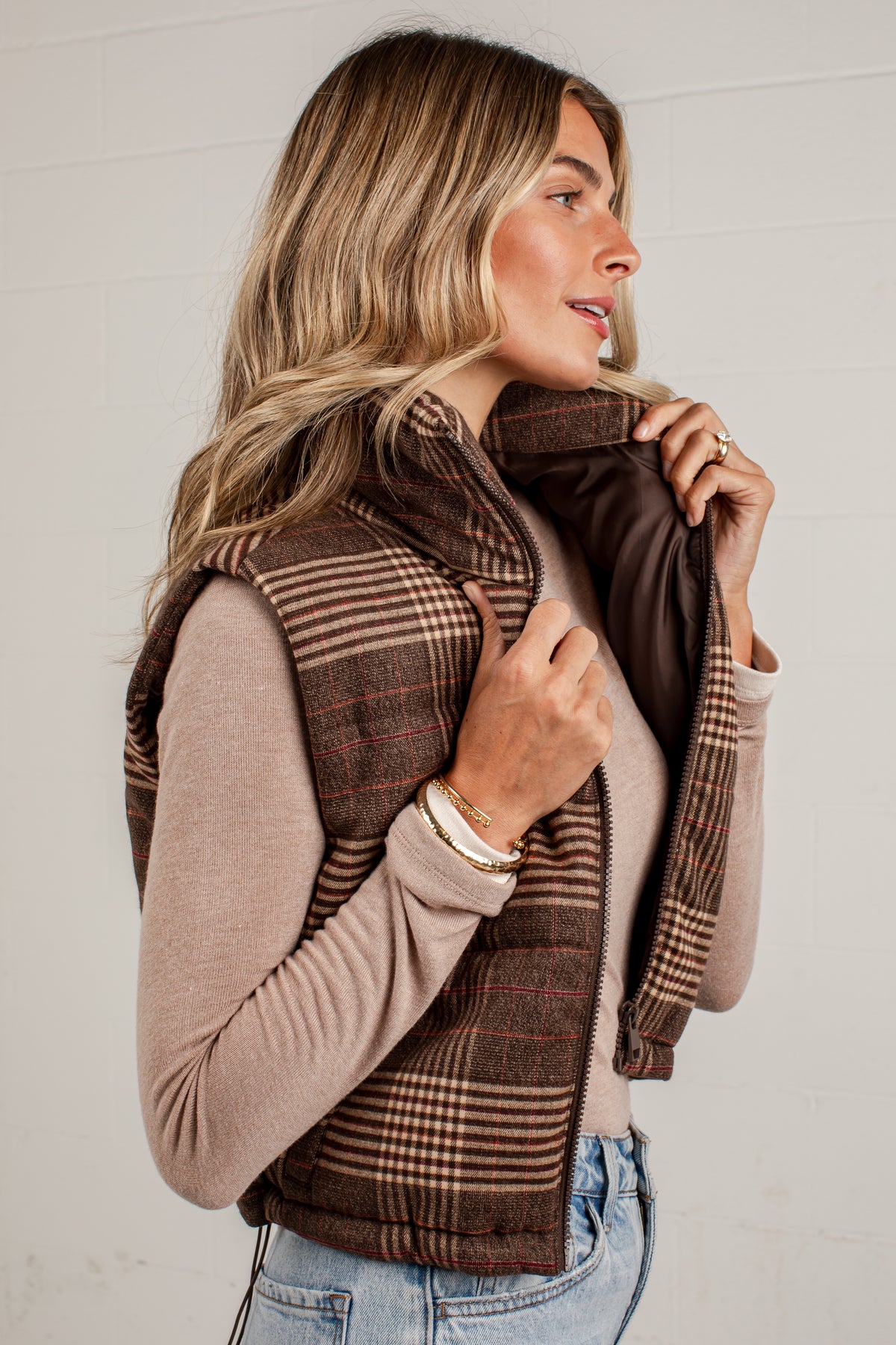 A woman wearing a brown plaid zippered vest over a beige long-sleeve top and light blue jeans, standing against a white paneled wall.
