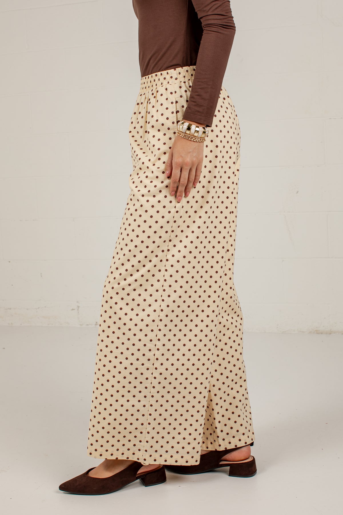 A person shown from the waist down wearing a cream-colored maxi skirt with a brown polka dot pattern, paired with a brown long-sleeve top and brown flat shoes, standing against a light off-white wall.