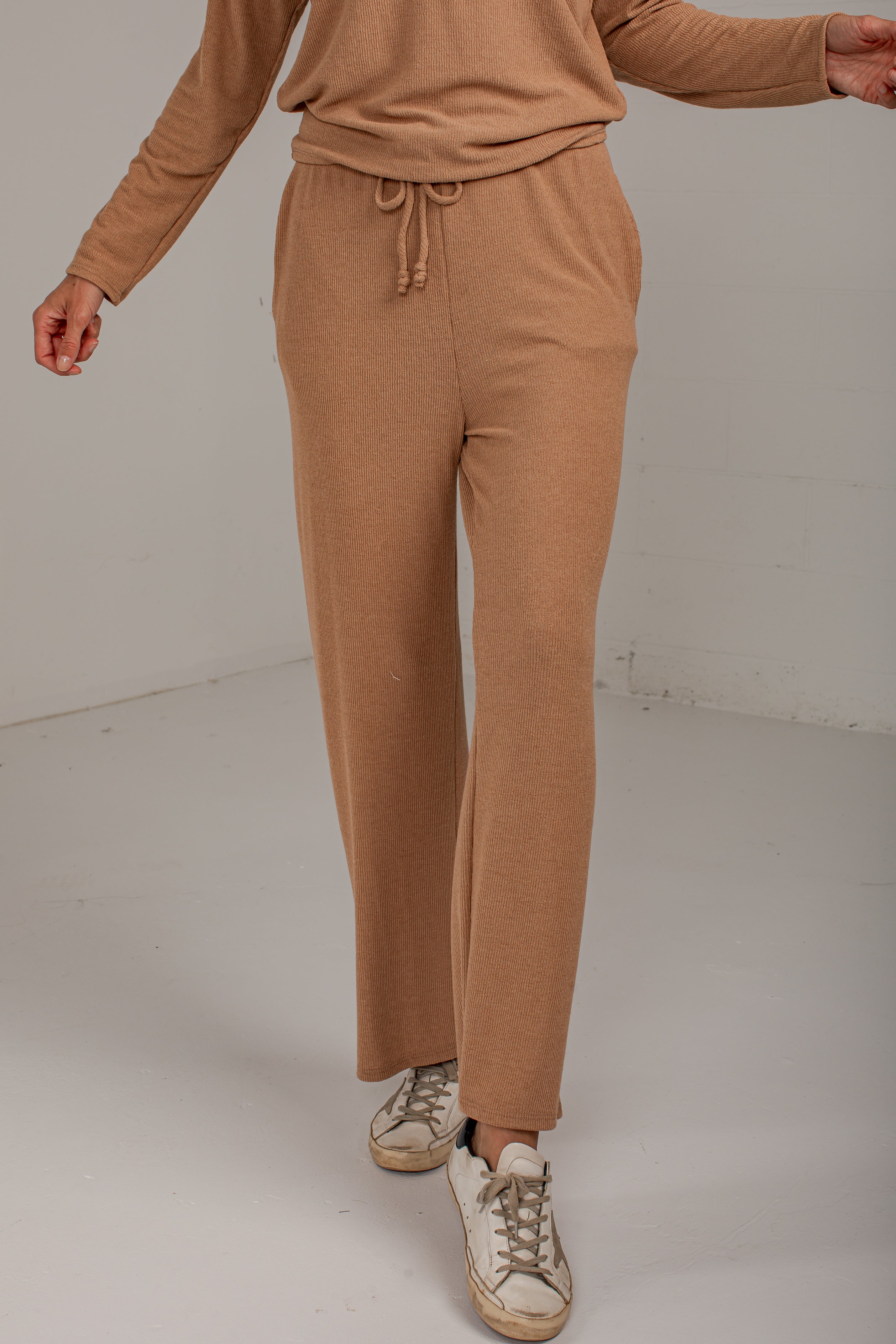 A person wearing a matching tan knit lounge set, featuring wide-leg drawstring pants and a long-sleeve top, paired with distressed white sneakers, photographed in a minimalist studio setting.