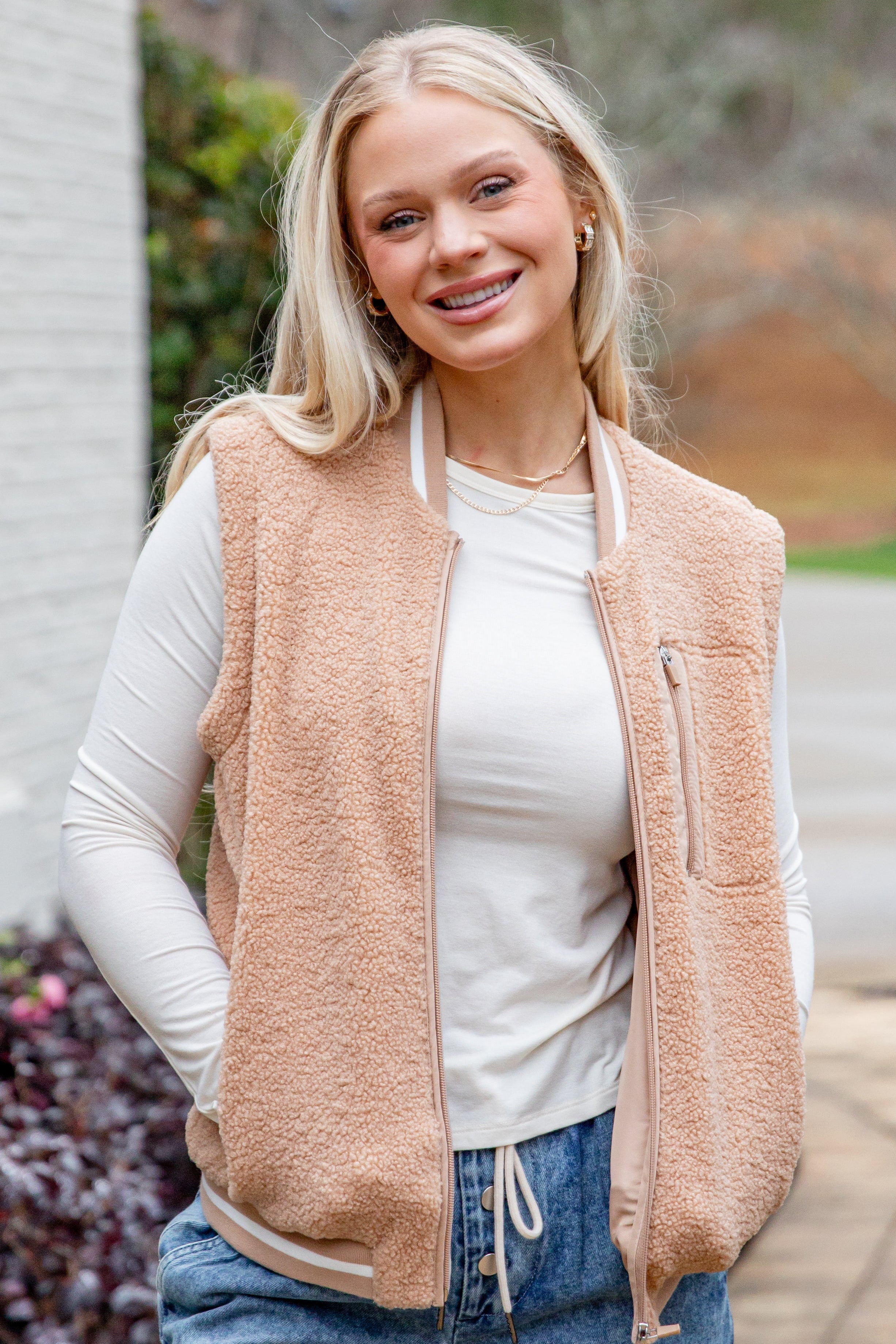 Woman wearing a beige vest over a white shirt with blue jeans, standing outdoors.